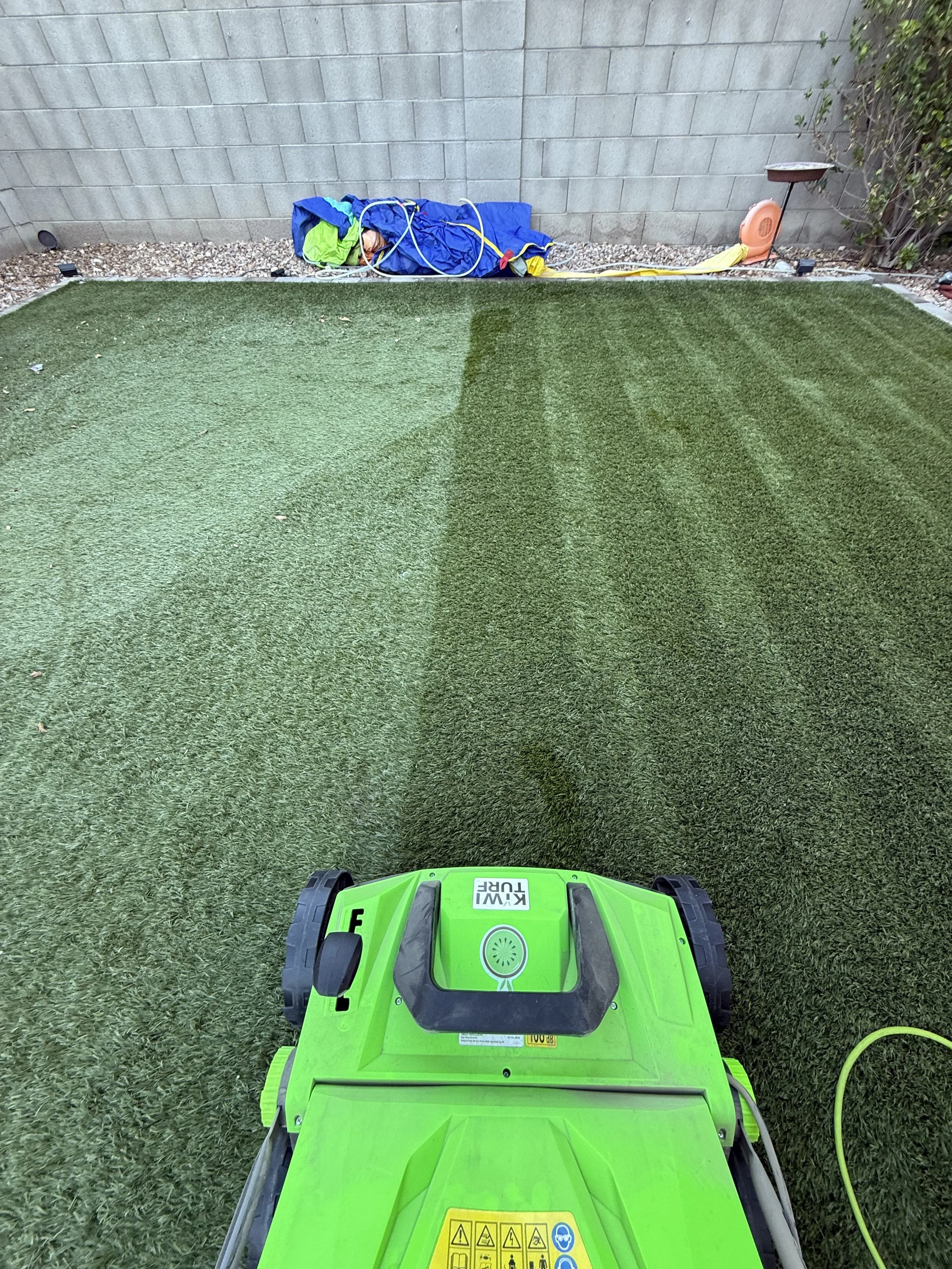 View from the operator's perspective of a green Kiwibot lawn mower on artificial grass, with a concrete block wall and pile of children’s clothes or fabric in the background, and a small electric fan on the right side.