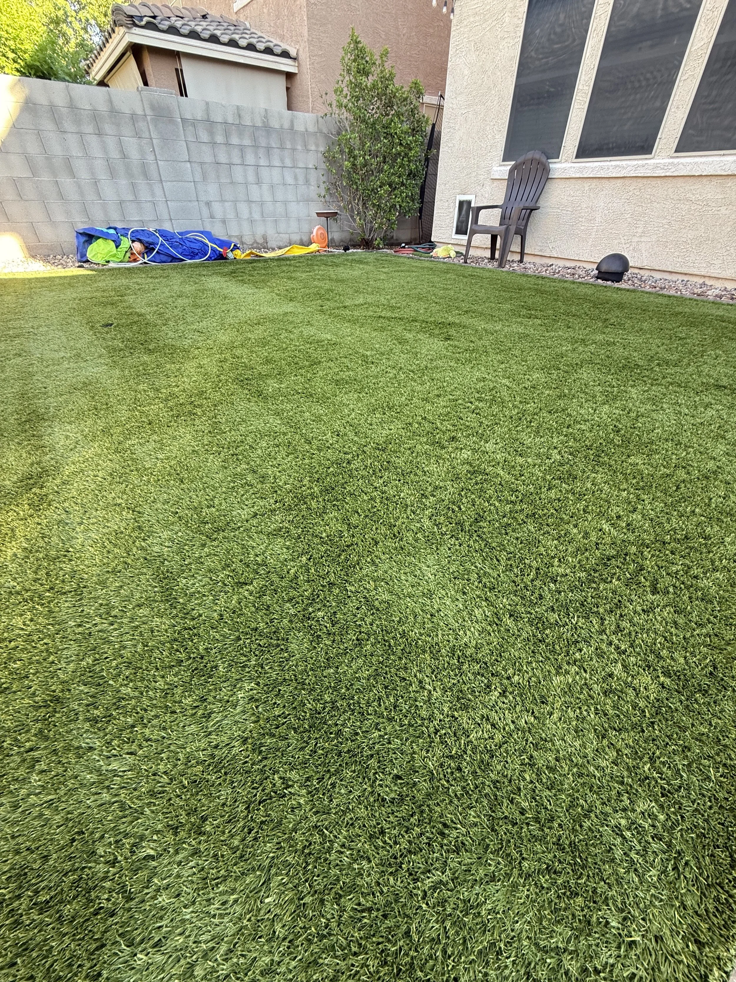 Lawn with artificial grass in a backyard, with a plastic chair, hedge, garden hose, and some tools against the house wall.