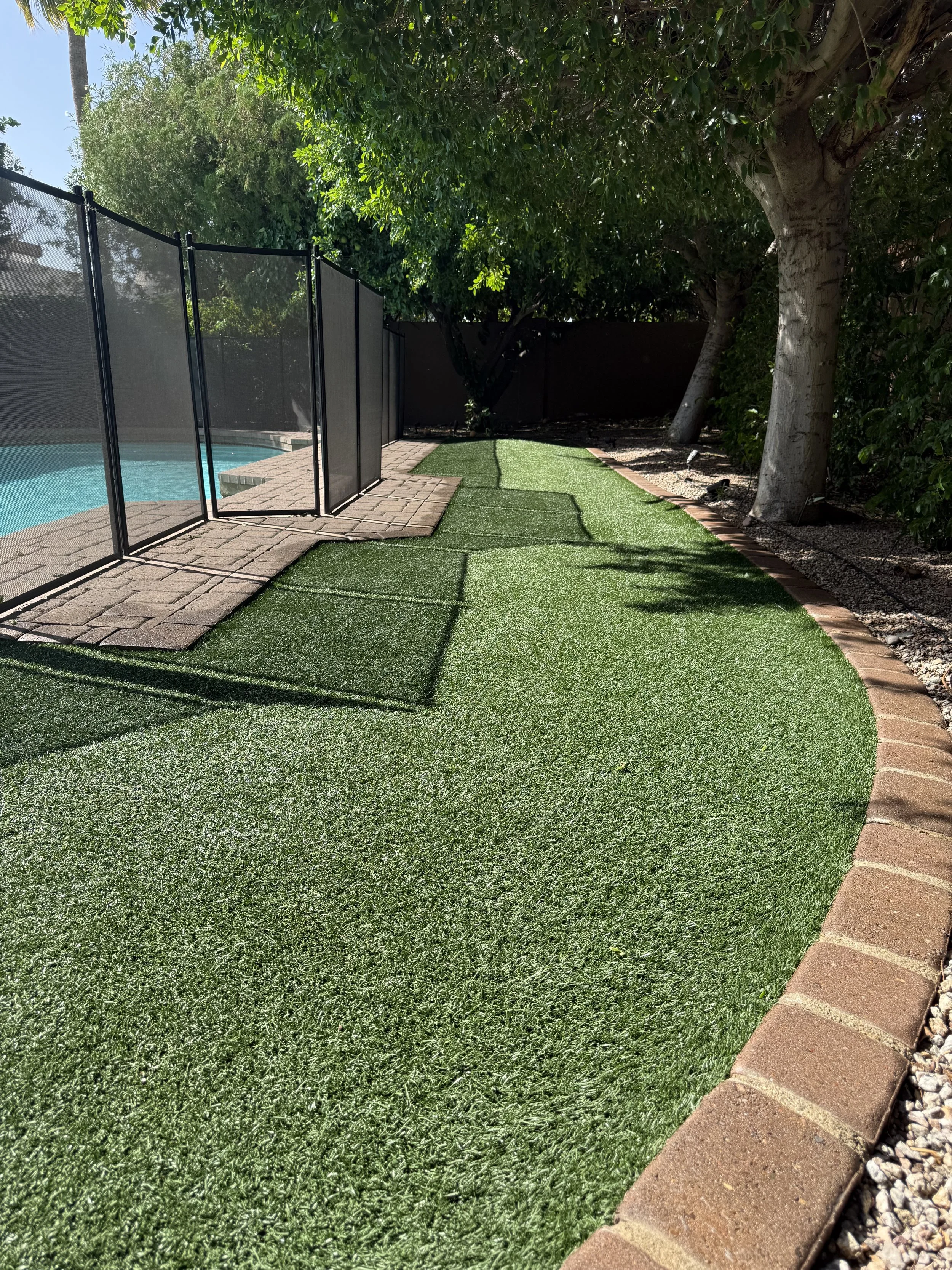 A backyard with artificial grass, a small swimming pool, a brick border, a brick pathway, a large tree, and a black privacy fence.