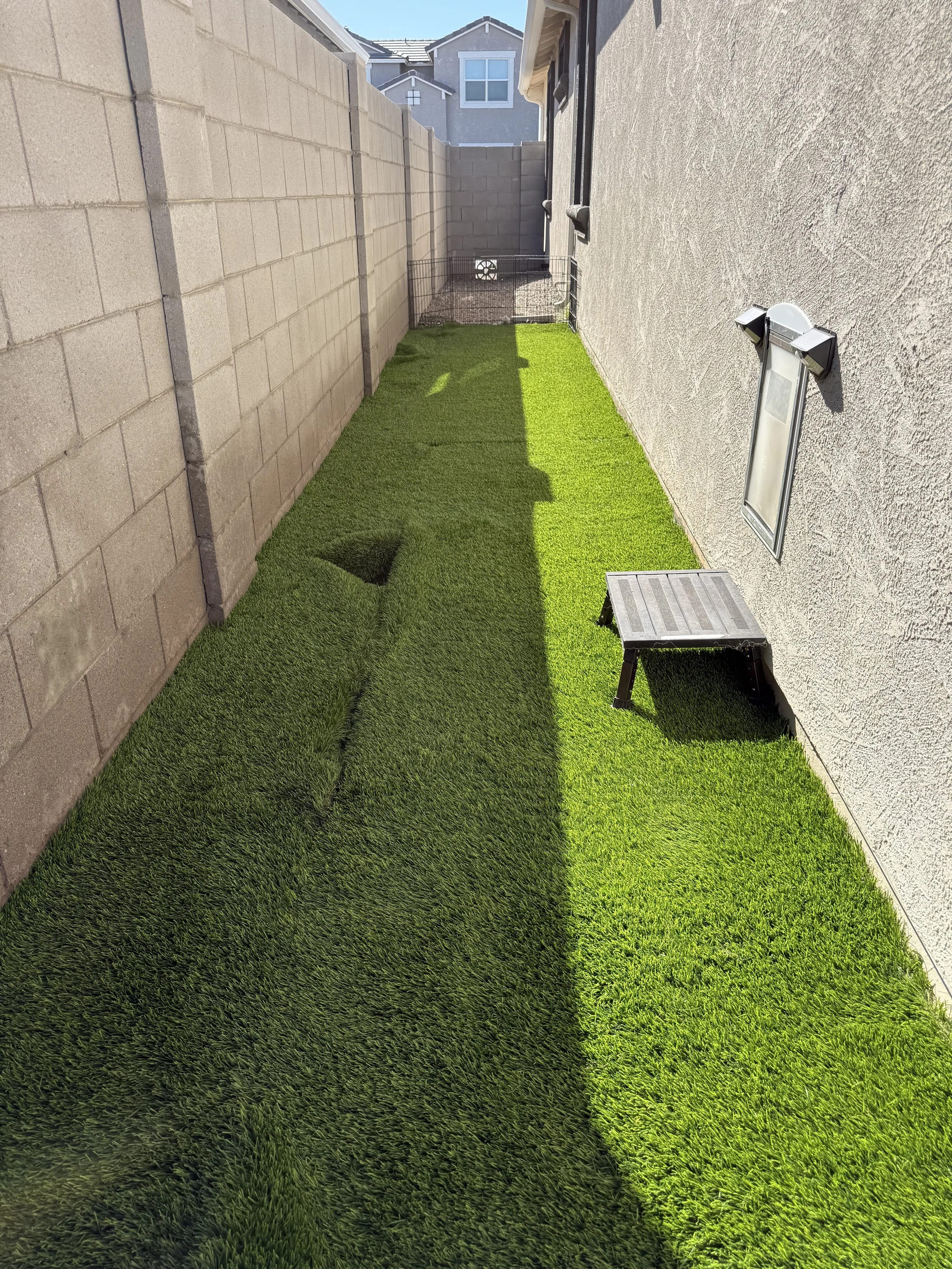 A narrow backyard with artificial grass, enclosed by a brick wall on the left and a beige stucco wall on the right, with a small window and outdoor light fixture visible.