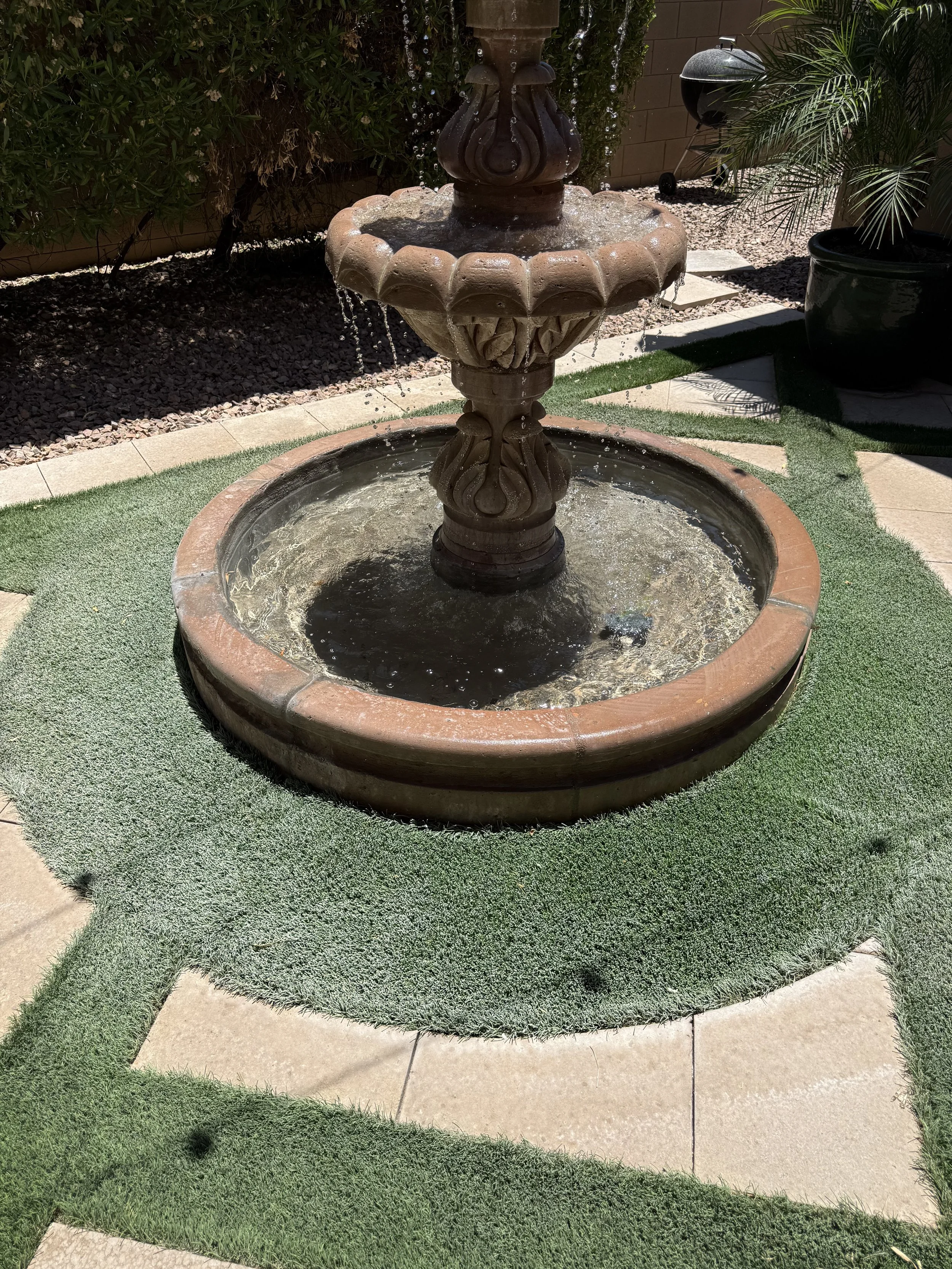 A two-tiered outdoor fountain with water flowing down, surrounded by a circular area with artificial grass and concrete tiles. There are plants and a barbecue grill in the background.