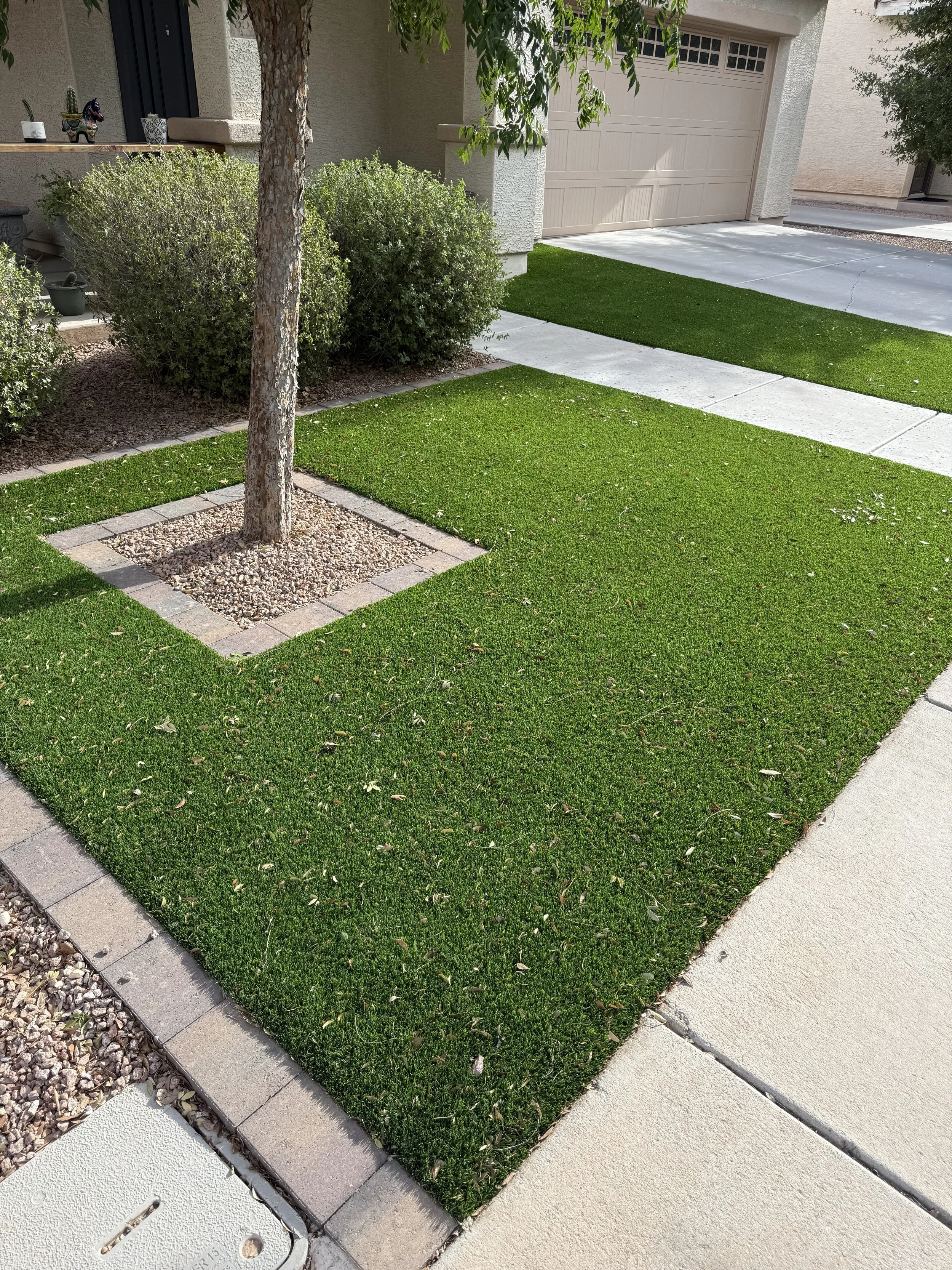 A front yard with a small tree, green grass patches bordered with bricks, a concrete sidewalk, and a yellow garage door.