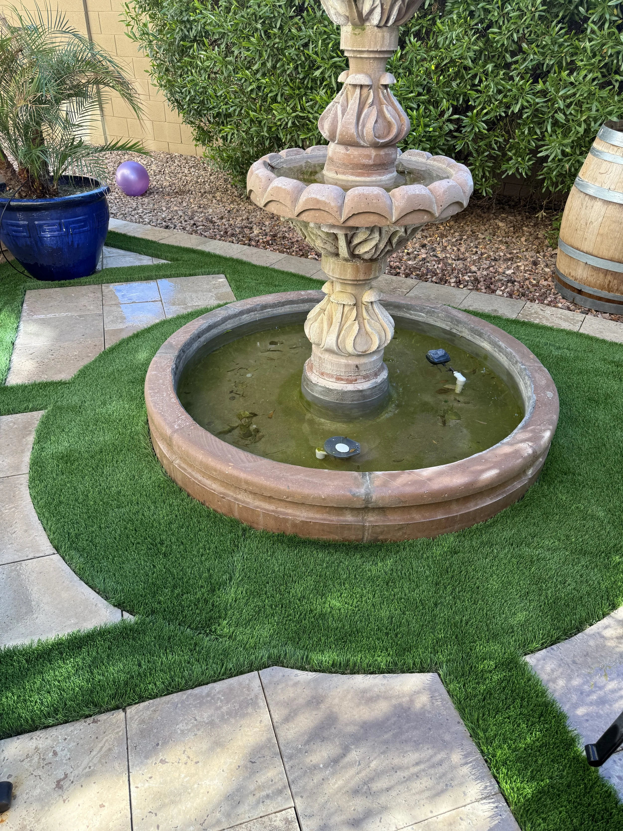 A stone fountain with water at the center of a backyard, surrounded by artificial green grass, concrete stepping stones, a potted plant, a decorative purple ball, and a large barrel in the background.