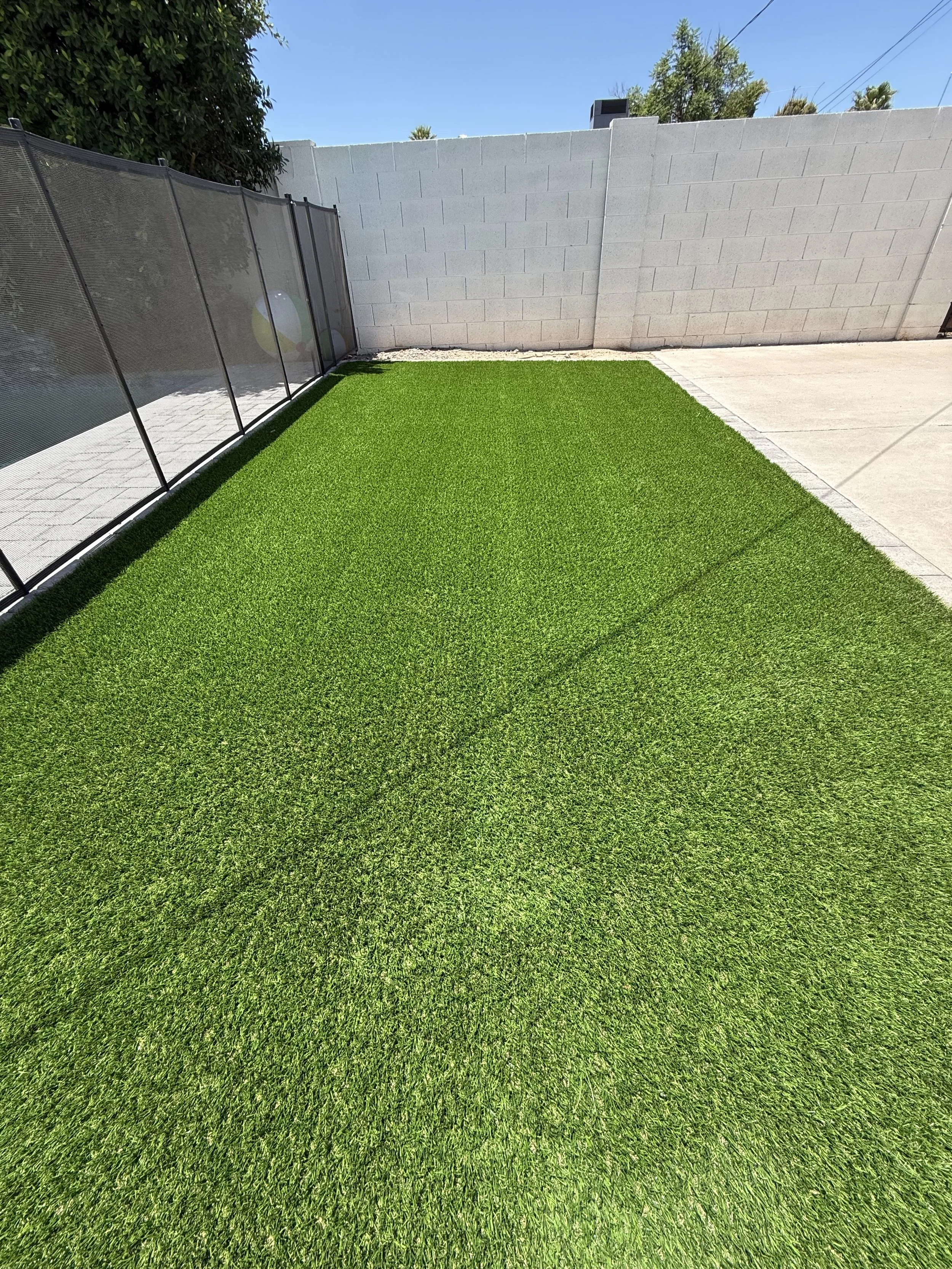 View of a backyard with a section of artificial green grass, concrete patio, a white cinder block wall, a black mesh fence, trees, and a blue sky.