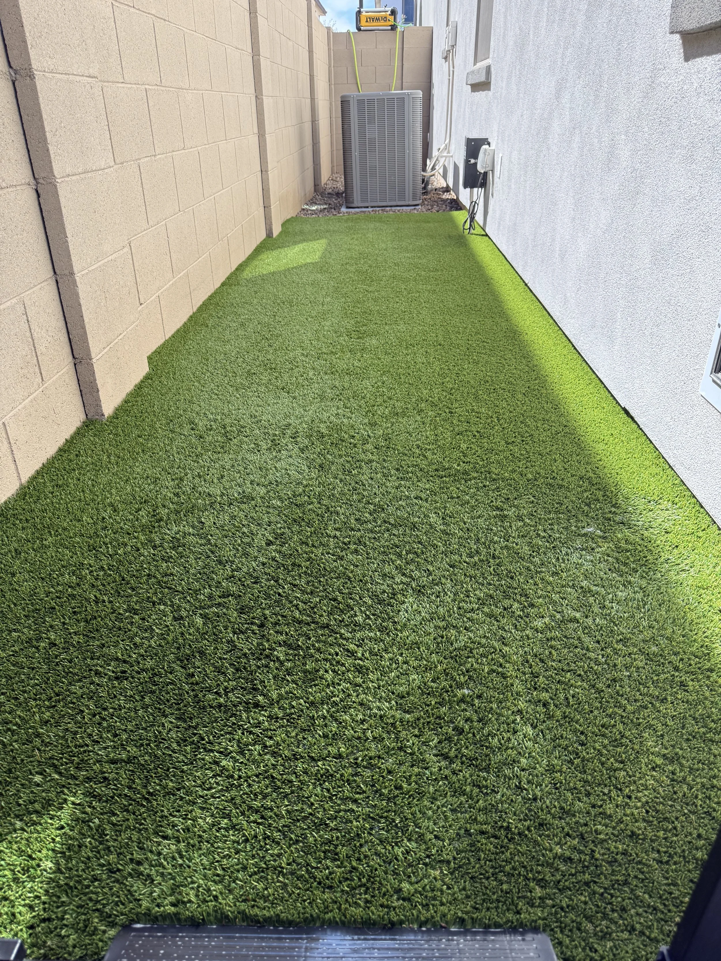A backyard with artificial green grass, beige brick wall on the left, gray stucco wall on the right, air conditioning unit at the back, and electrical equipment mounted on the stucco wall.