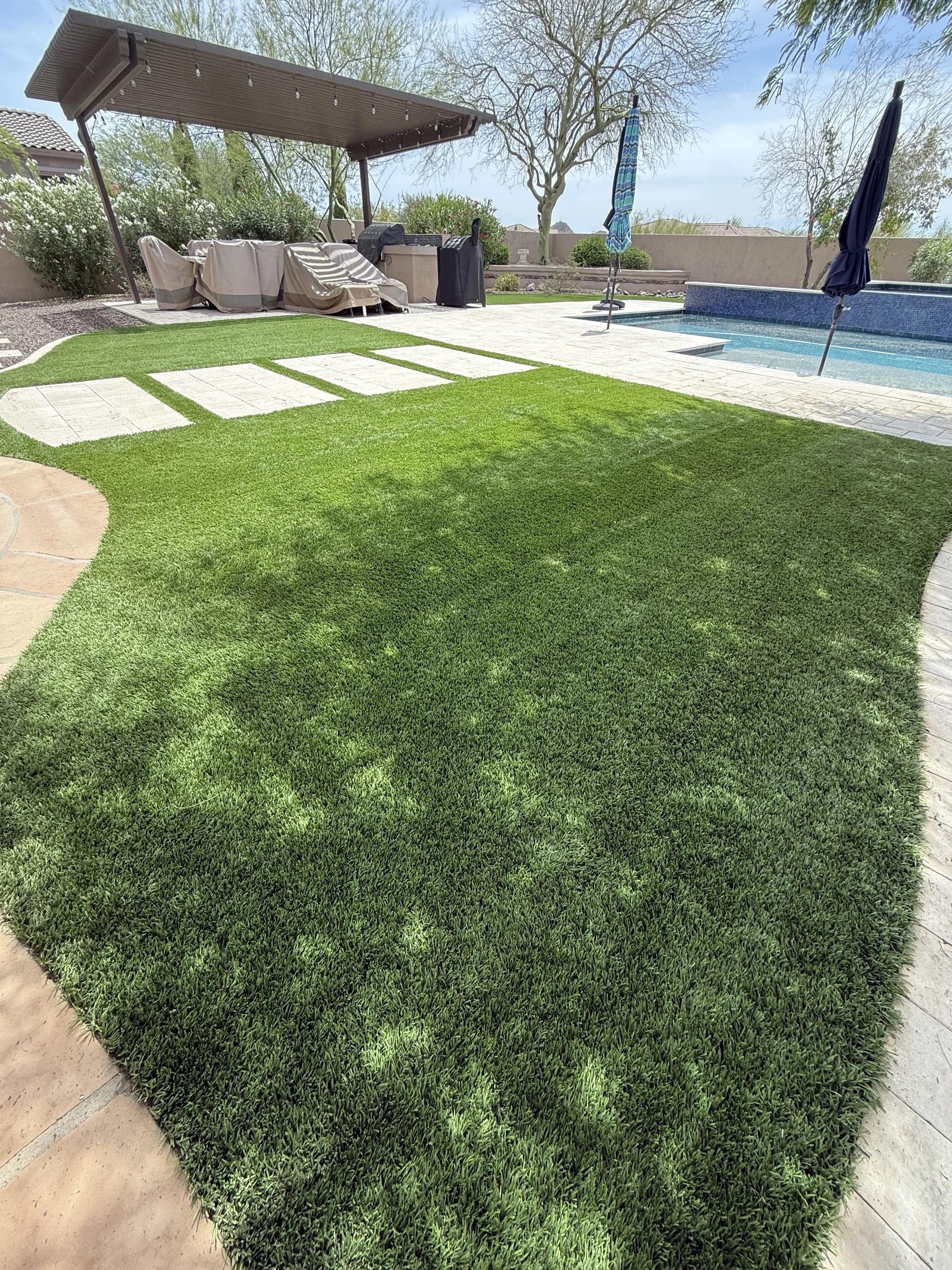 Backyard with well-manicured green grass, stone pavers, a pool with umbrellas, 2 covered outdoor furniture sets, a shaded seating area under a pergola, trees, and a concrete wall in the background.