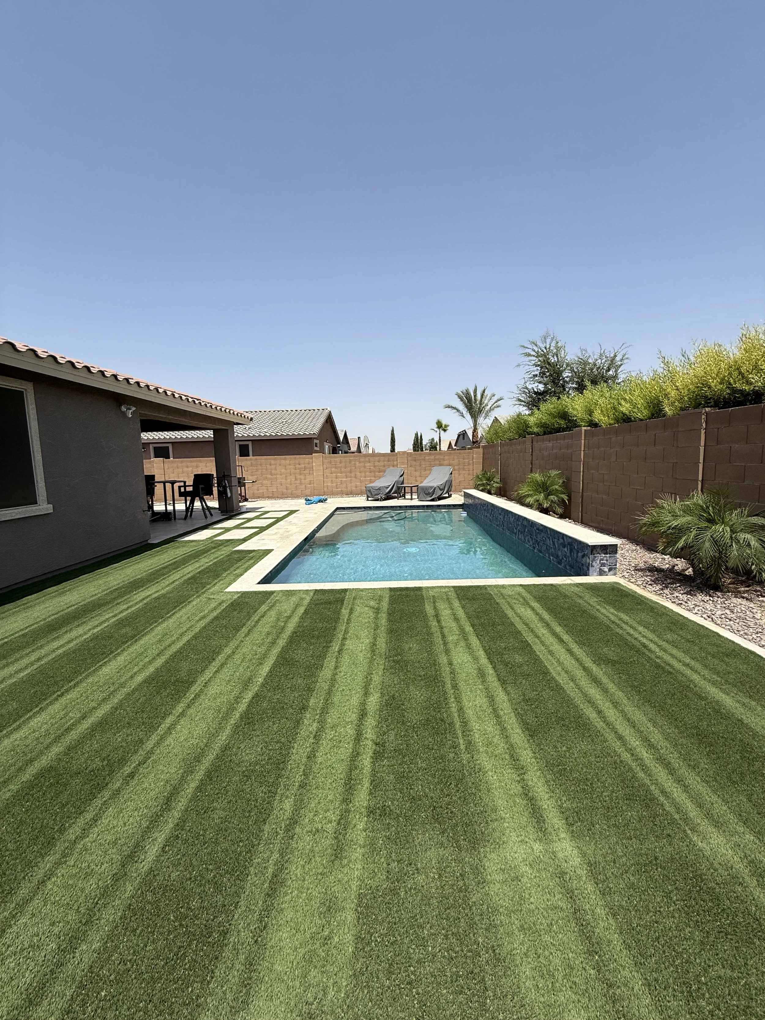 Backyard with a swimming pool, two lounge chairs, a covered patio area with outdoor seating, and a green artificial grass lawn, enclosed by a brick wall and desert plants under a clear blue sky.