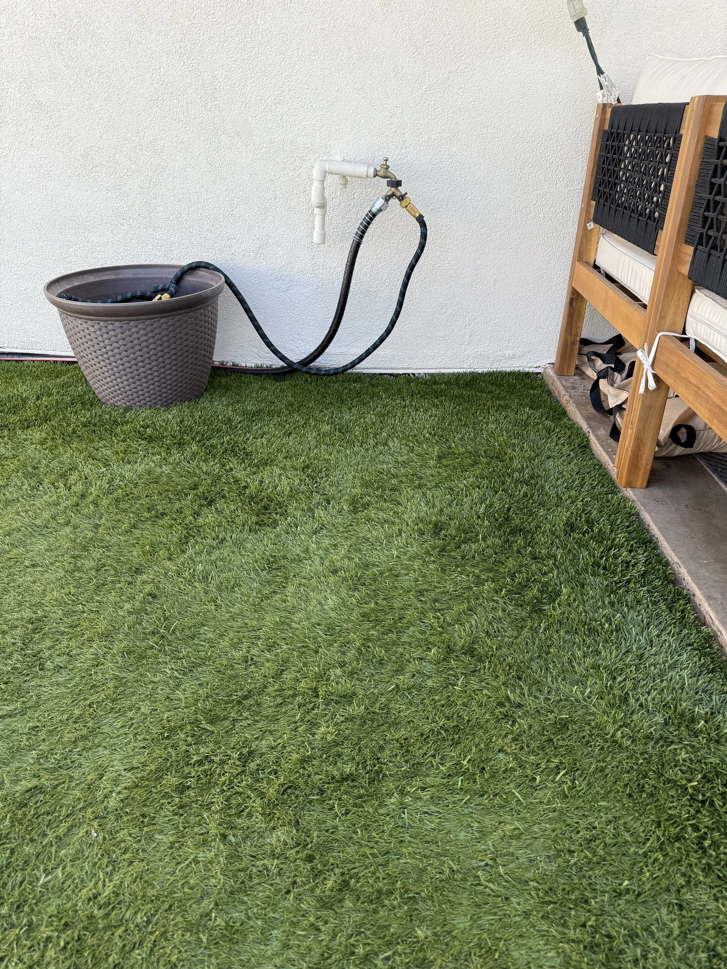 A garden hose connected to water fixture outside a house, with a black hose leading to a large patterned gray planter on green grass, next to a wooden outdoor bench.