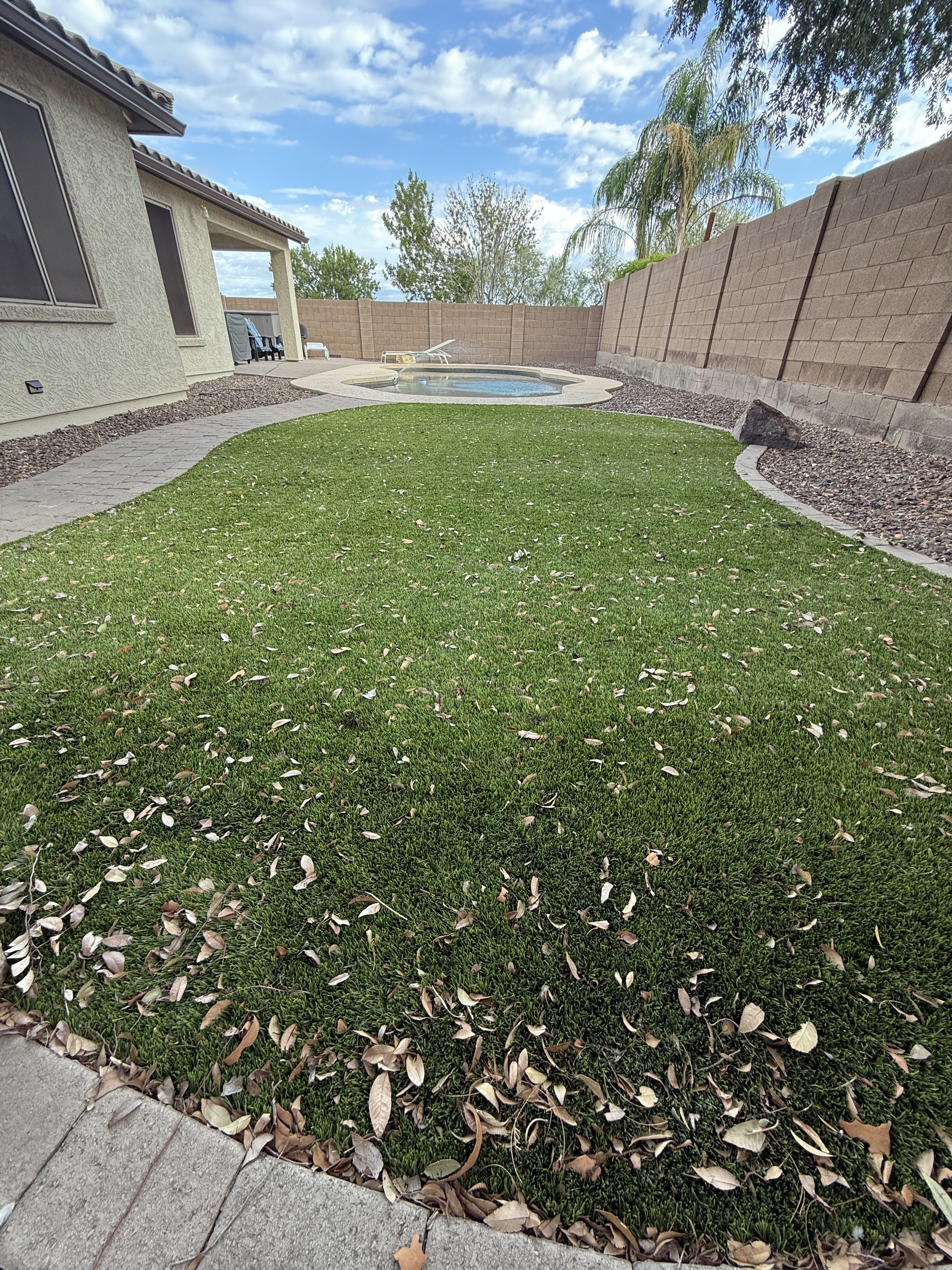 A backyard with a house on the left, a small pool, a green lawn, a stone walkway, a wooden fence, and trees under a partly cloudy sky.