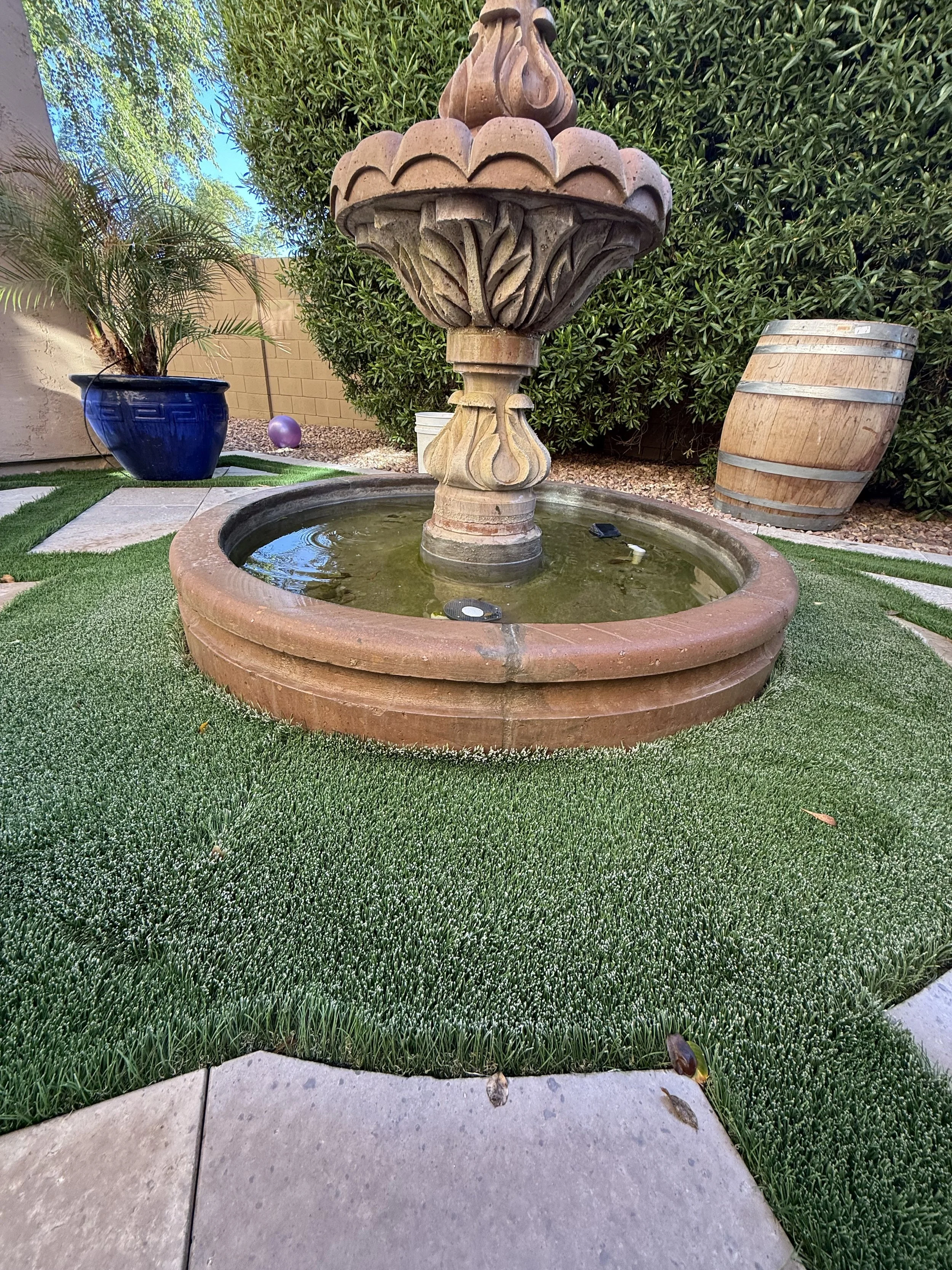 A backyard fountain with a carved stone top, surrounded by green grass, potted plants, mulch, and a wooden barrel on its side.