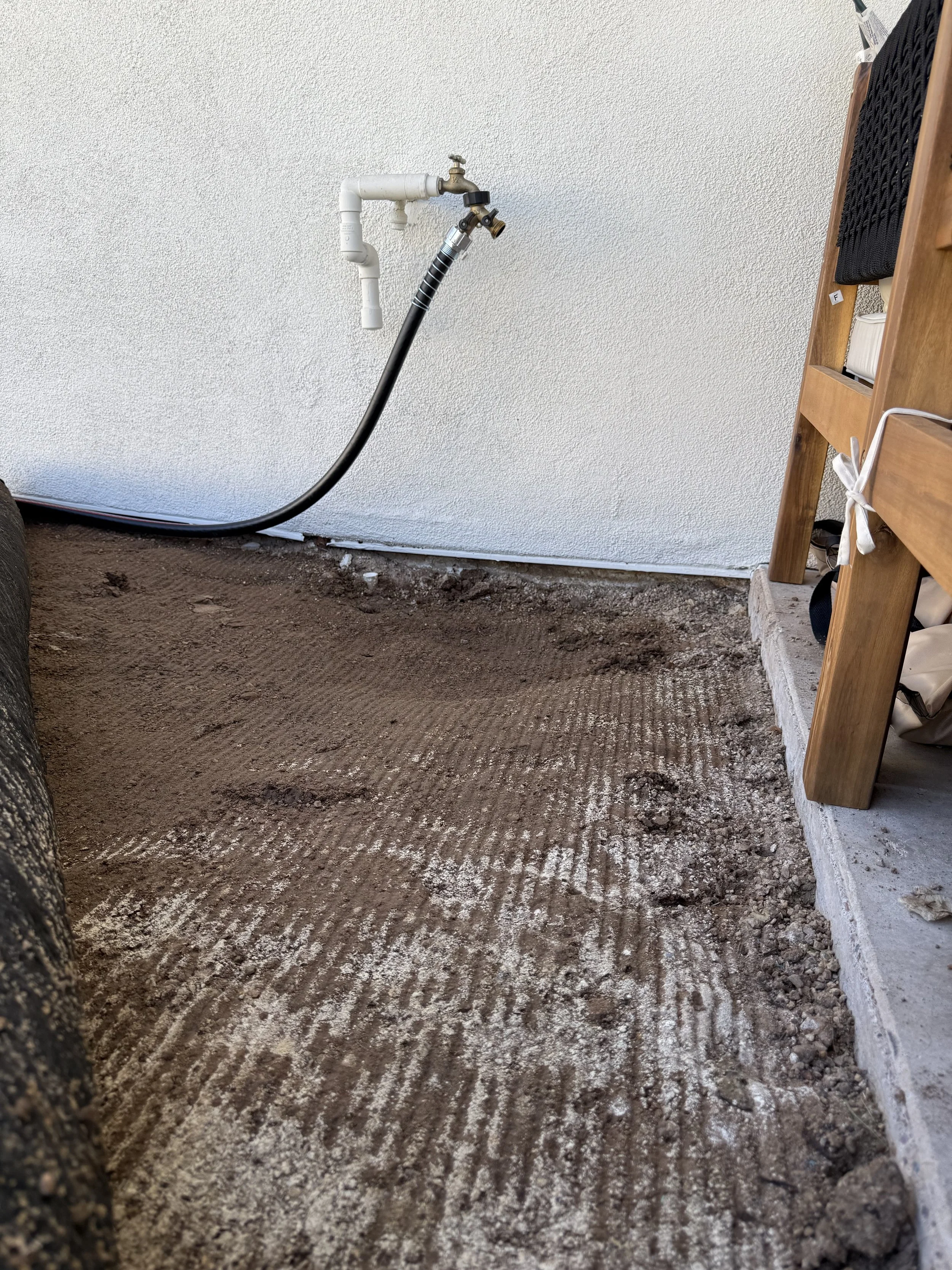 Close-up of a dirt floor with a partially dug trench next to a wall. There is a black hose connected to white pipes on the wall. A wooden piece of furniture with a black mat and a white bag is visible on the right side.