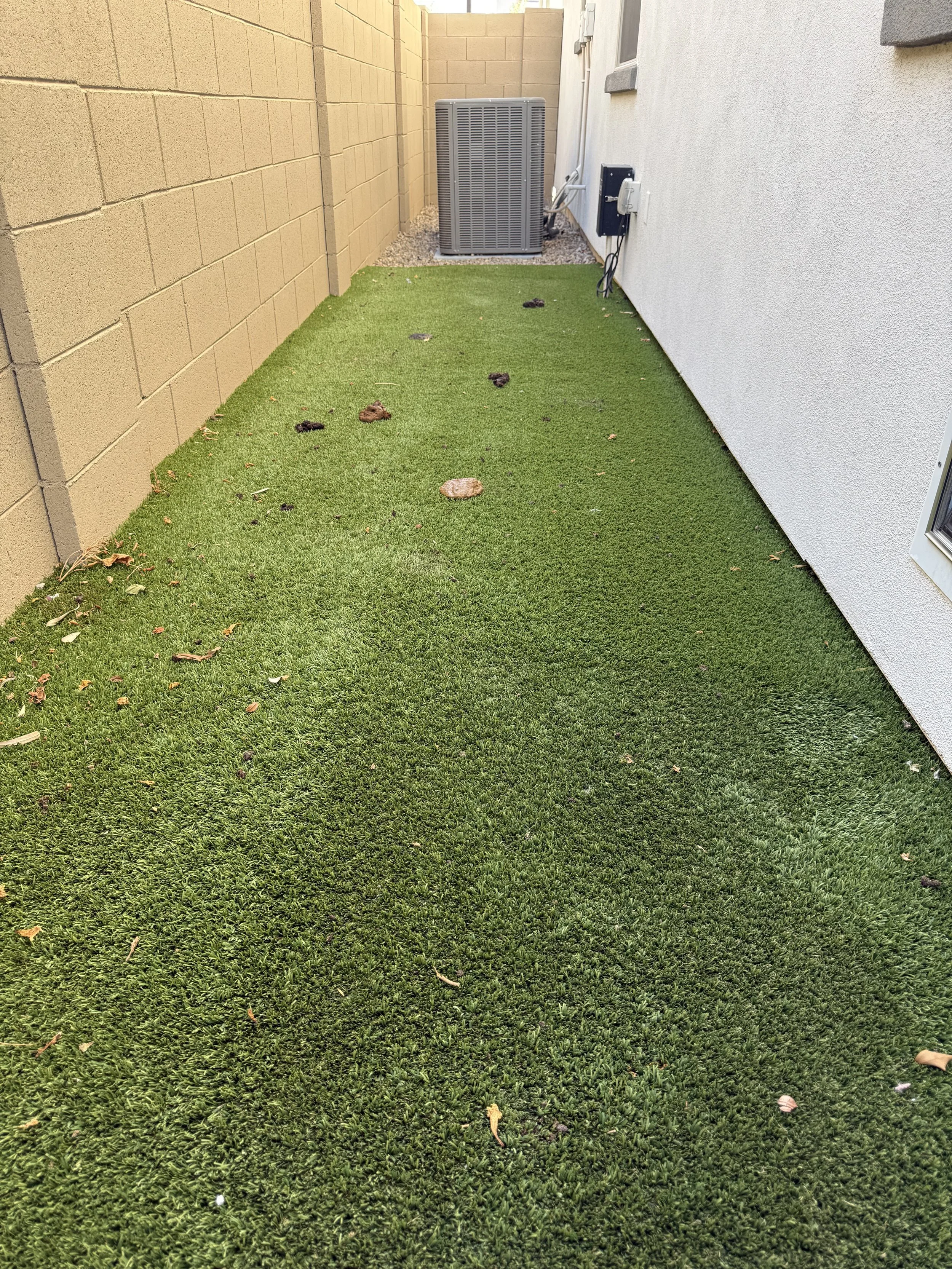 Backyard with artificial grass, beige brick wall on the left, and white exterior wall on the right. An air conditioning unit and electrical box are visible near the back, with some scattered debris on the grass.