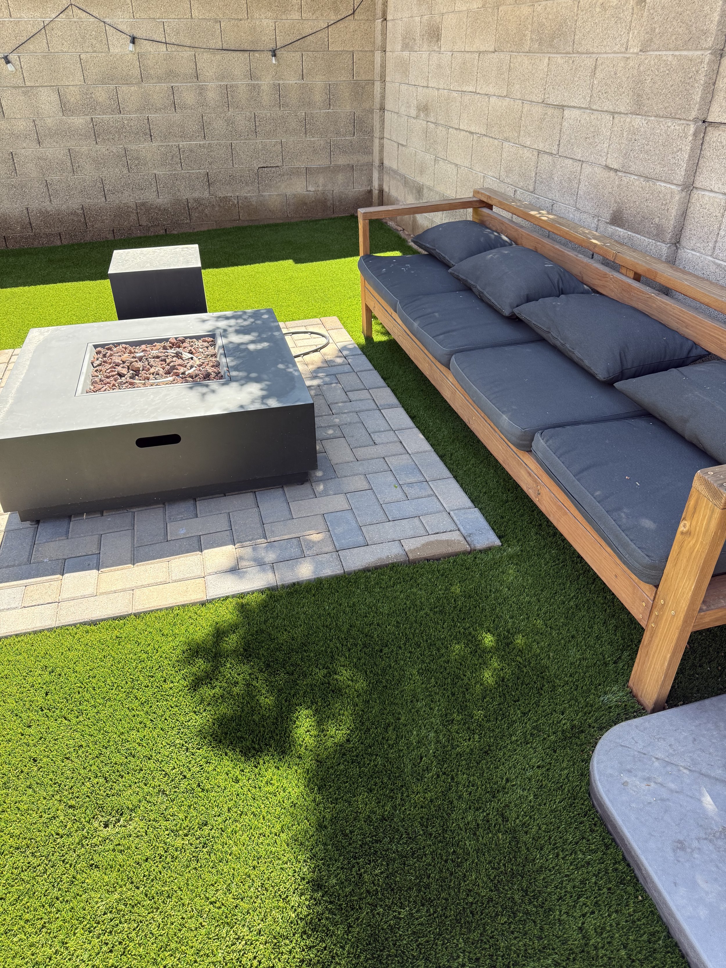 A backyard patio area with artificial grass, a stone fire pit, a wooden bench with black cushions, and a cinder block wall with string lights above.