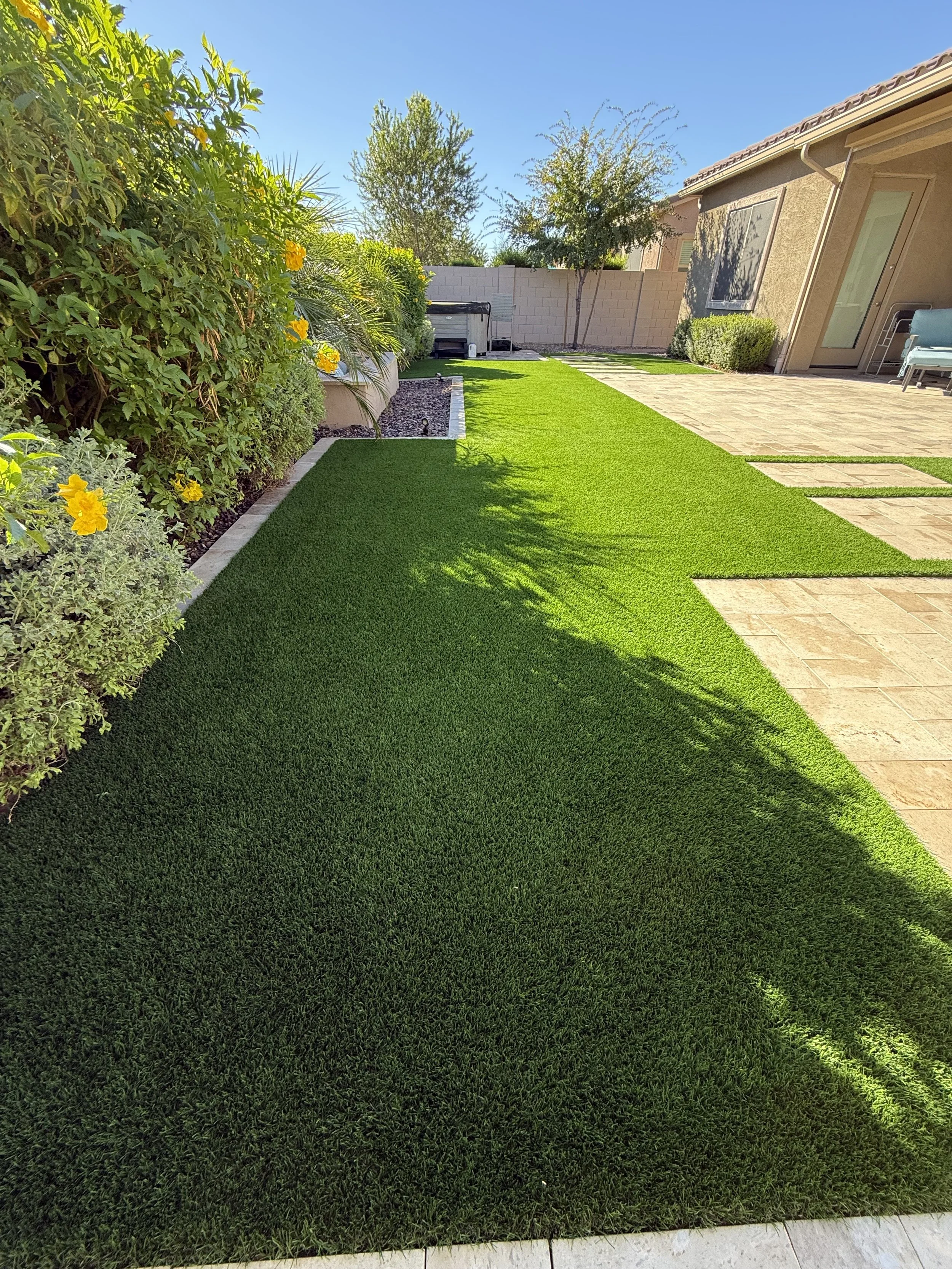 A backyard with a well-maintained green lawn, bordered by colorful flowering bushes and trees, with a brick patio area and a house with a sliding glass door on the right side.