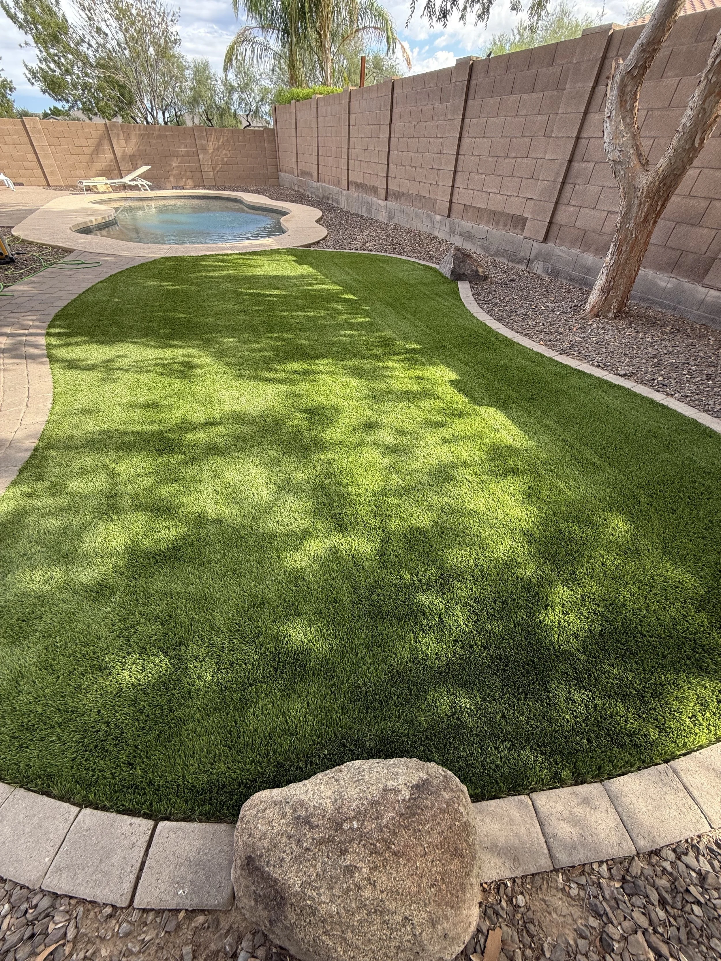 Backyard with a small swimming pool, artificial grass, a stone border, and a tall brick fence, with a large rock near the edge of the grass.