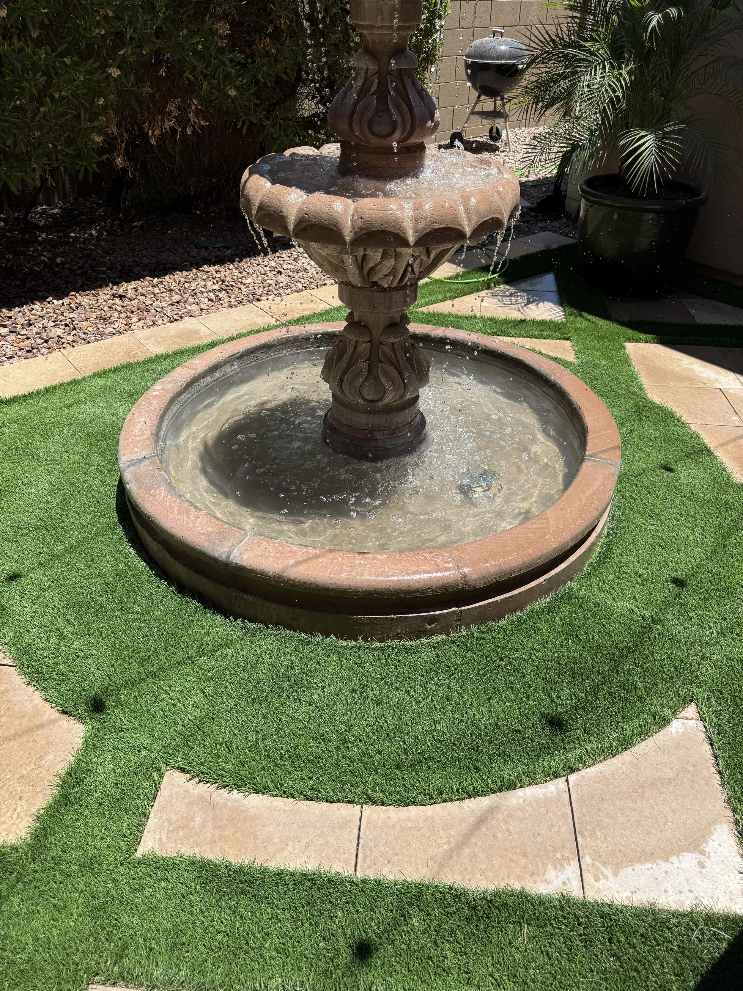 A decorative outdoor fountain with water flowing from multiple tiers, surrounded by artificial grass with a paved walkway nearby. In the background, there are plants, a gravel bed, and a small barbecue grill.