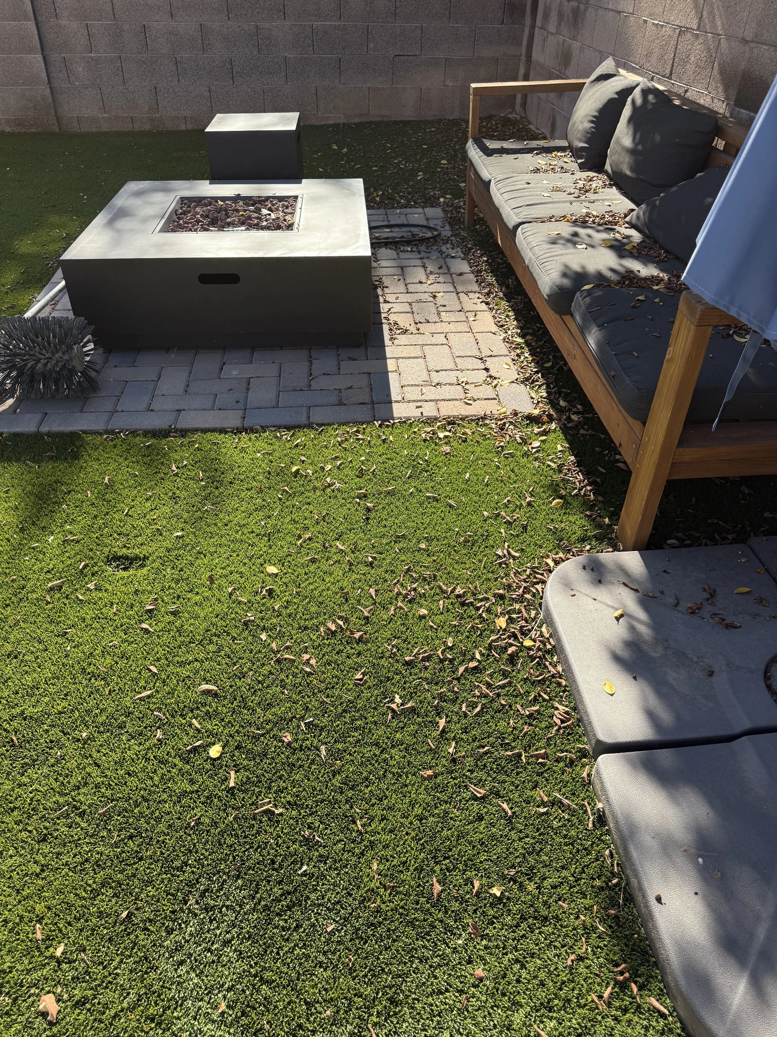 An outdoor backyard scene with a green lawn, a brick patio, a fire pit filled with rocks, a gray bench with black cushions and a pillow, and a bouncy brush on the grass.