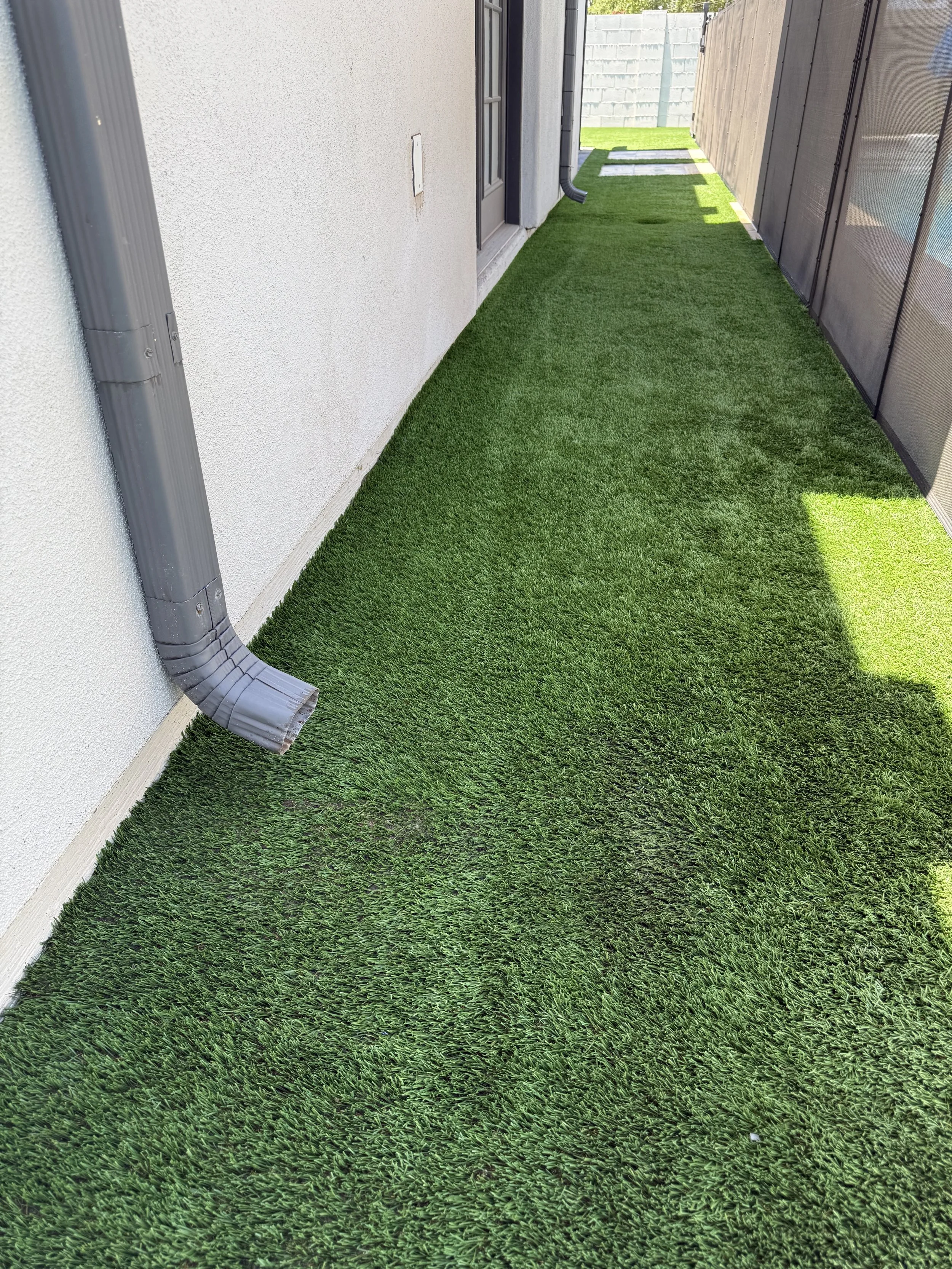 An outdoor side yard with artificial green grass, a white exterior wall on the left, a wooden fence on the right, and a grey downspout extending from the roof to the ground. There is a door with a screen and a window behind it, with a section of gras