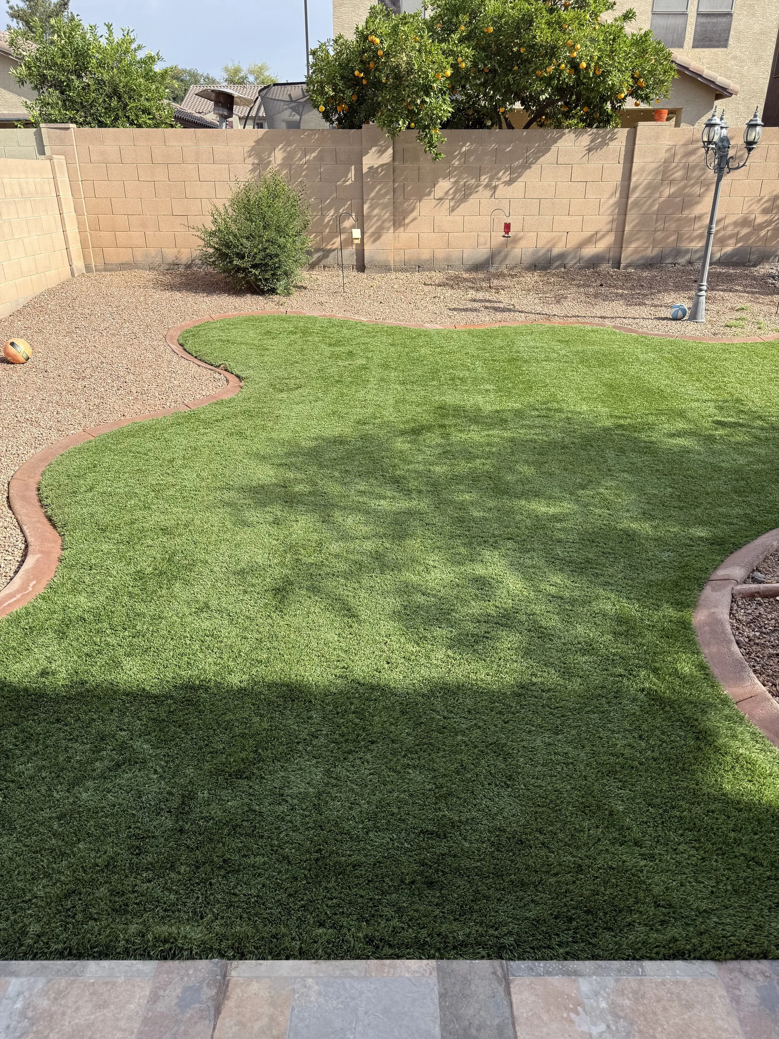 A backyard scene with a patch of green grass bordered by a stone walkway, a brick wall, and a gravel area. There are trees and plants, including an orange tree, and a decorative lamp post with three lanterns.