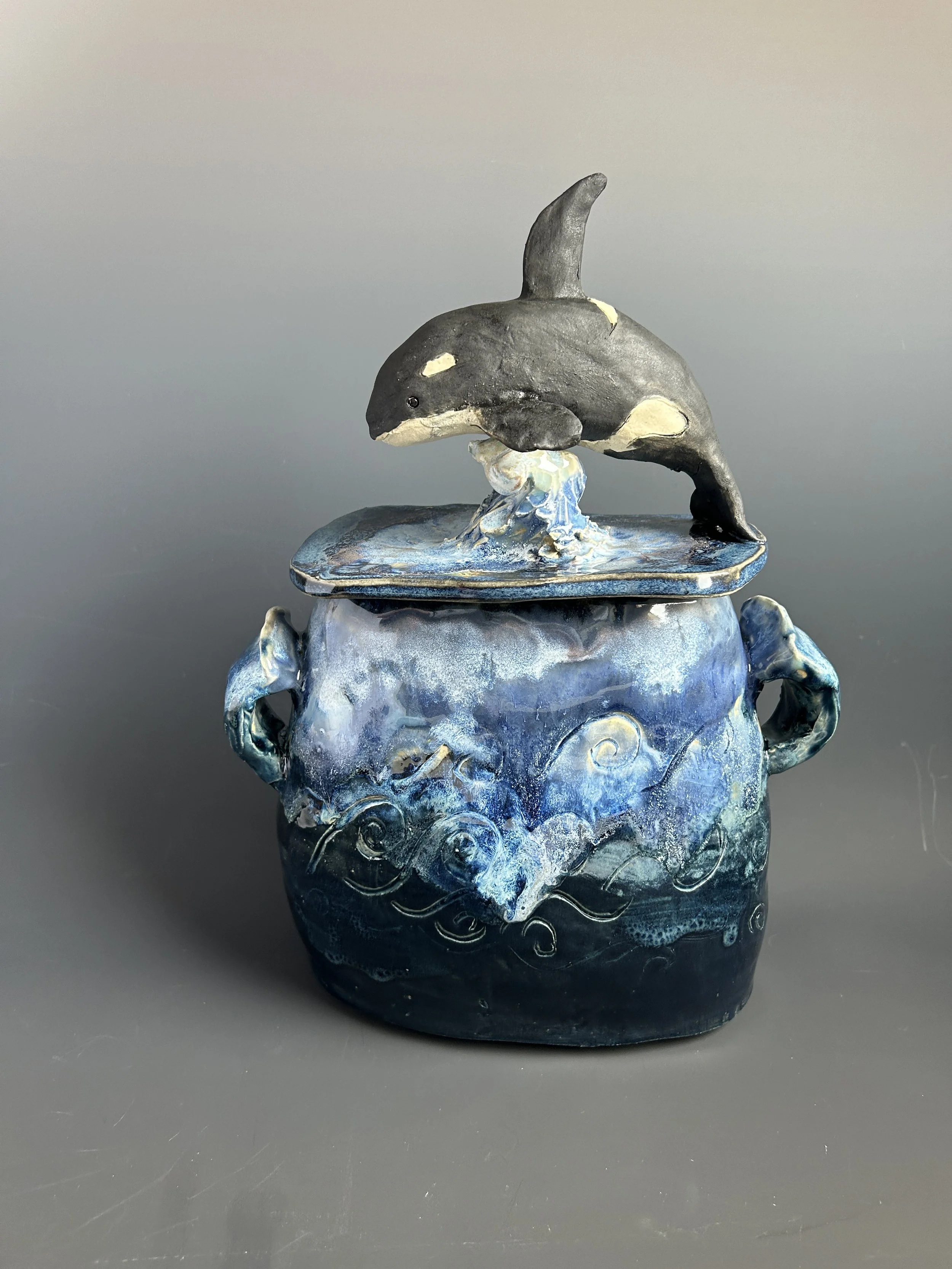 Handmade ceramic jar of a whale jumping out of water, mounted on a blue and black painted wooden base with swirling wave patterns.