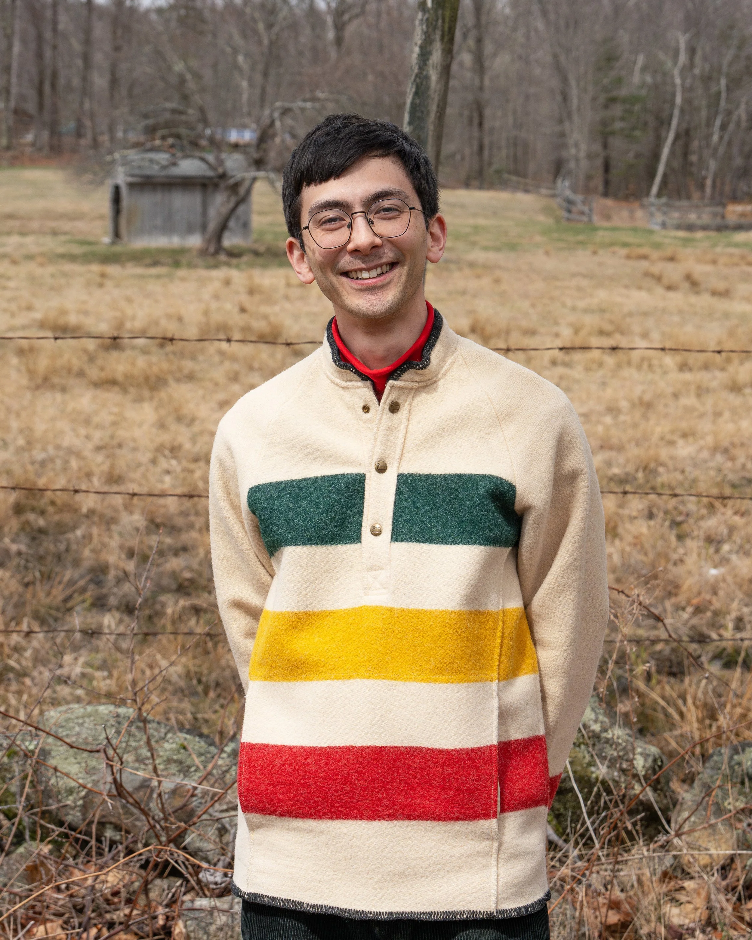 A young man with glasses and a gray T-shirt smiling outdoors in a lush green natural setting.