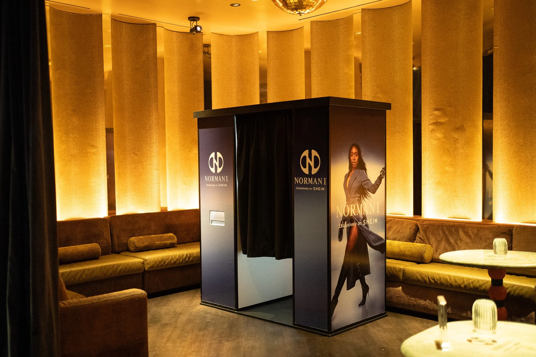 A vintage photo booth setup outside against a white wall, with a black curtain and a wooden body on a paved sidewalk, surrounded by bushes and part of a brick wall on the left.