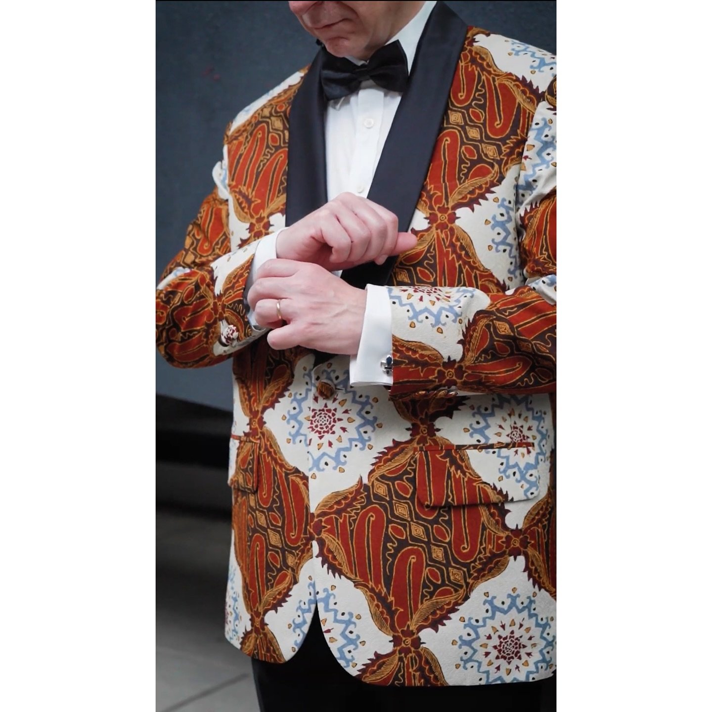 A bold fusion of tradition and sophistication &mdash; this striking tuxedo features a rich batik-inspired pattern in deep crimson, gold, and chestnut tones, subtly echoing the vibrant hues of the Spanish flag. Set against a soft cream background, the