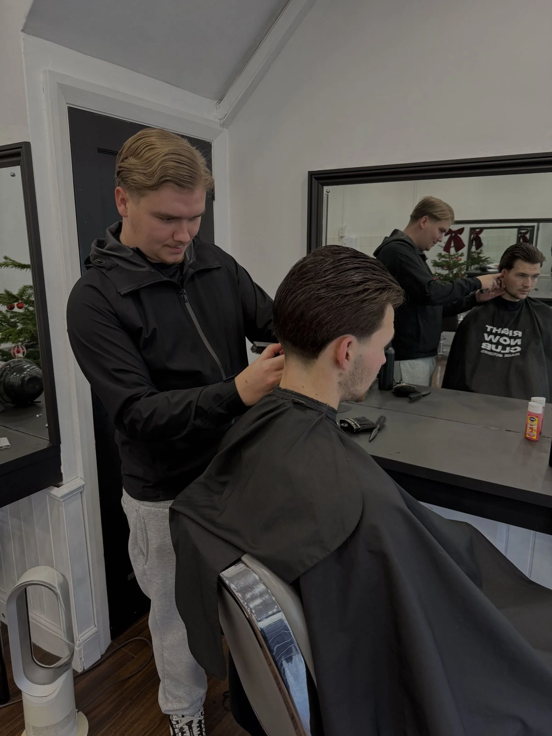 Barbershop with a barber giving a haircut to a male customer seated in a chair. The barber is standing and using scissors to trim the hair. There is a mirror behind the customer reflecting the barber and the shop's interior, including Christmas decorations on the window.