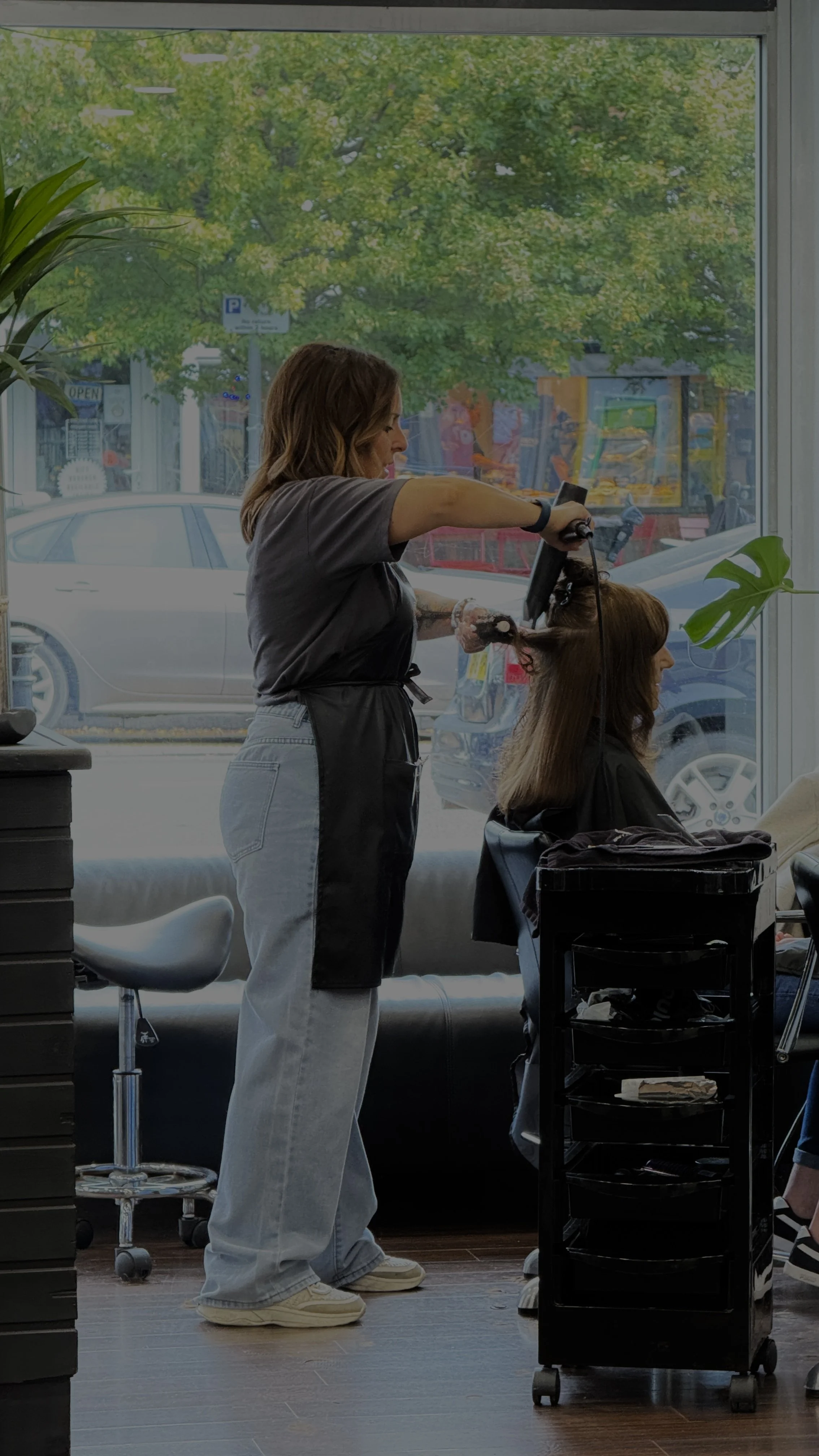 A woman sitting on a chair at a salon inside, with hair stylist standing behind her, and a large window showing an outside view of buildings, trees, a red mailbox, and parked cars.