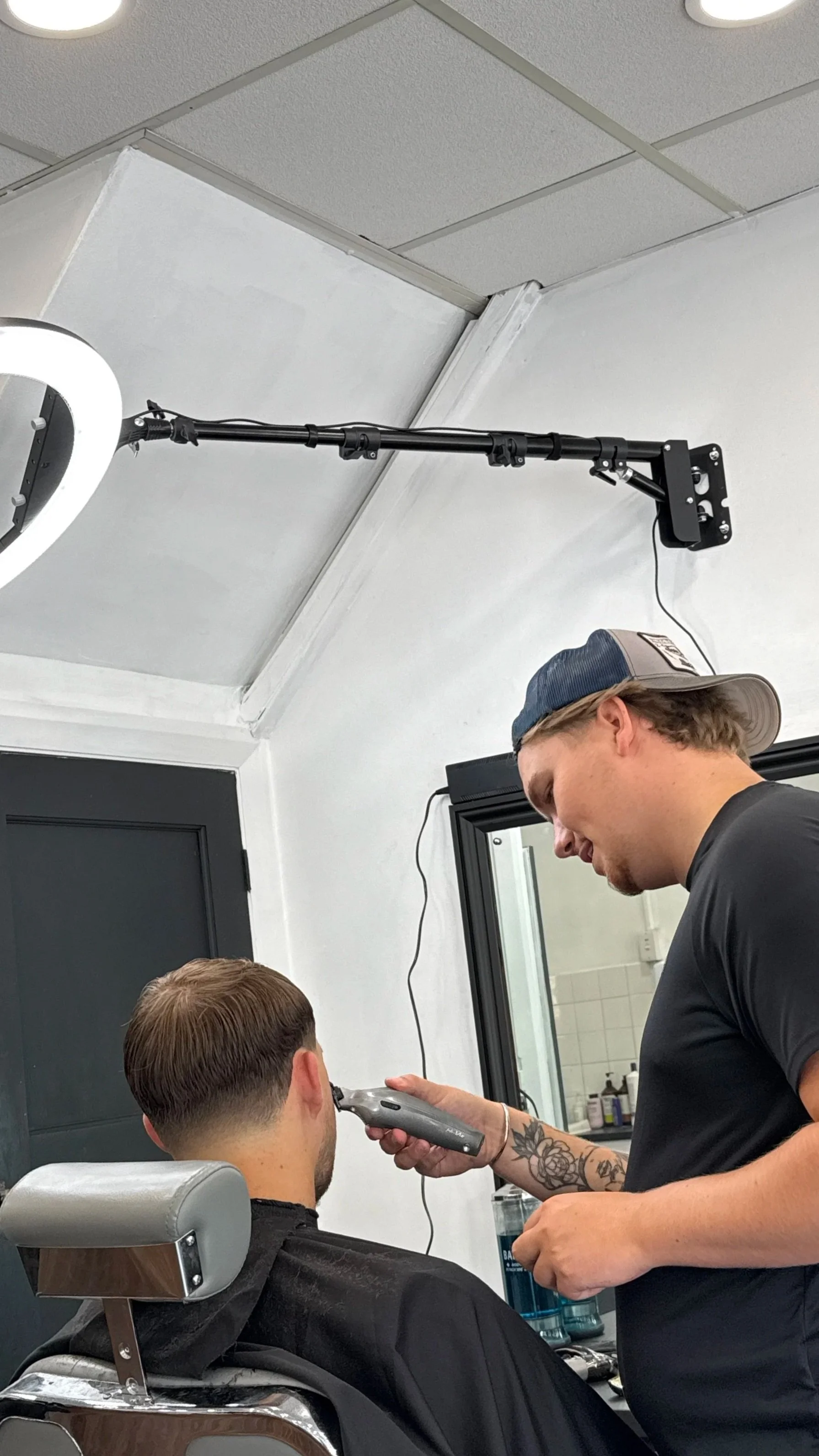 Barbershop with a barber giving a haircut to a male customer seated in a chair. The barber is standing and using scissors to trim the hair. There is a mirror behind the customer reflecting the barber and the shop's interior, including Christmas decorations on the window.