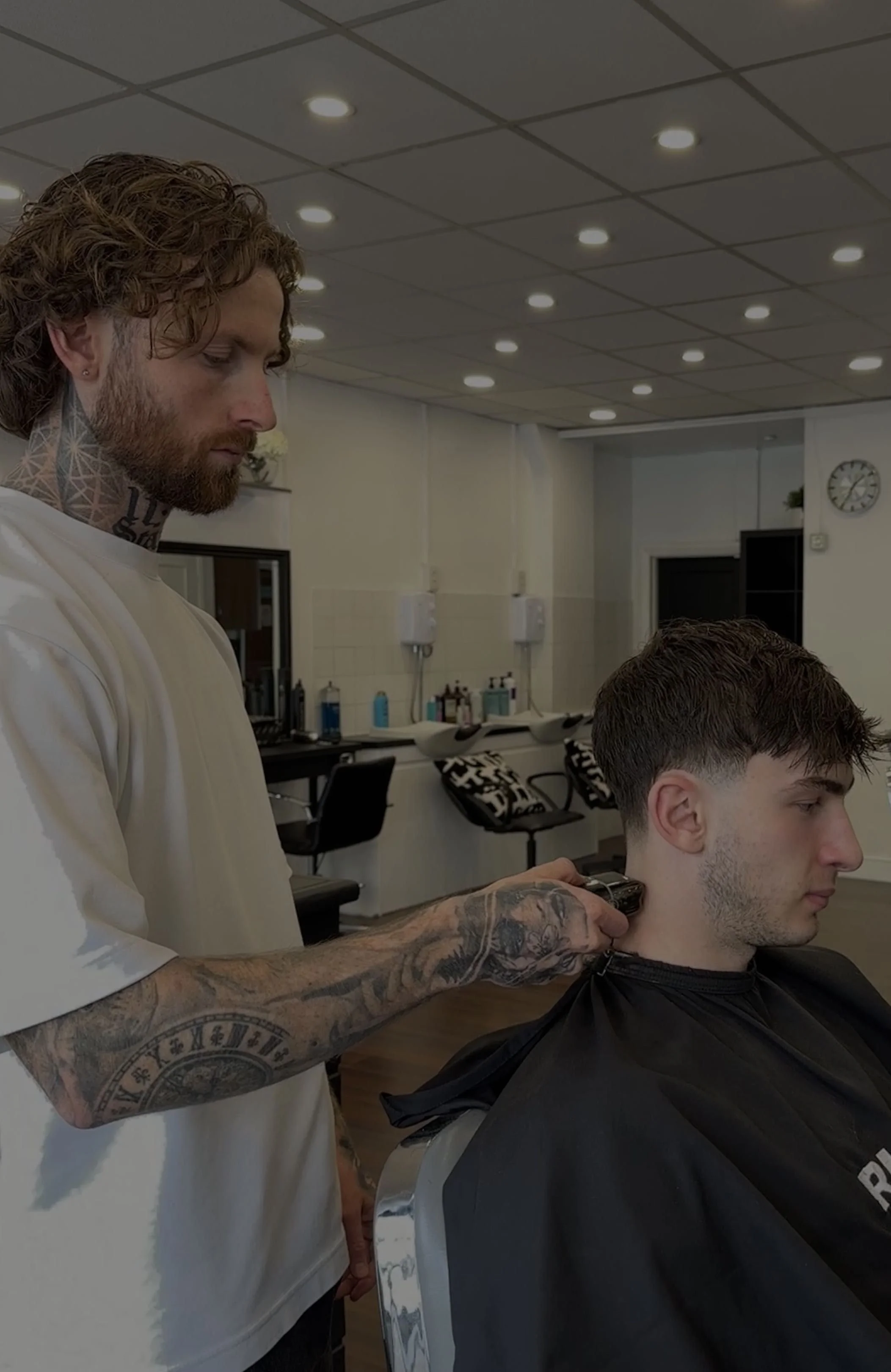 Barber trimming a young man's hair in a barbershop with white walls, ceiling lights, and various products on the counters.
