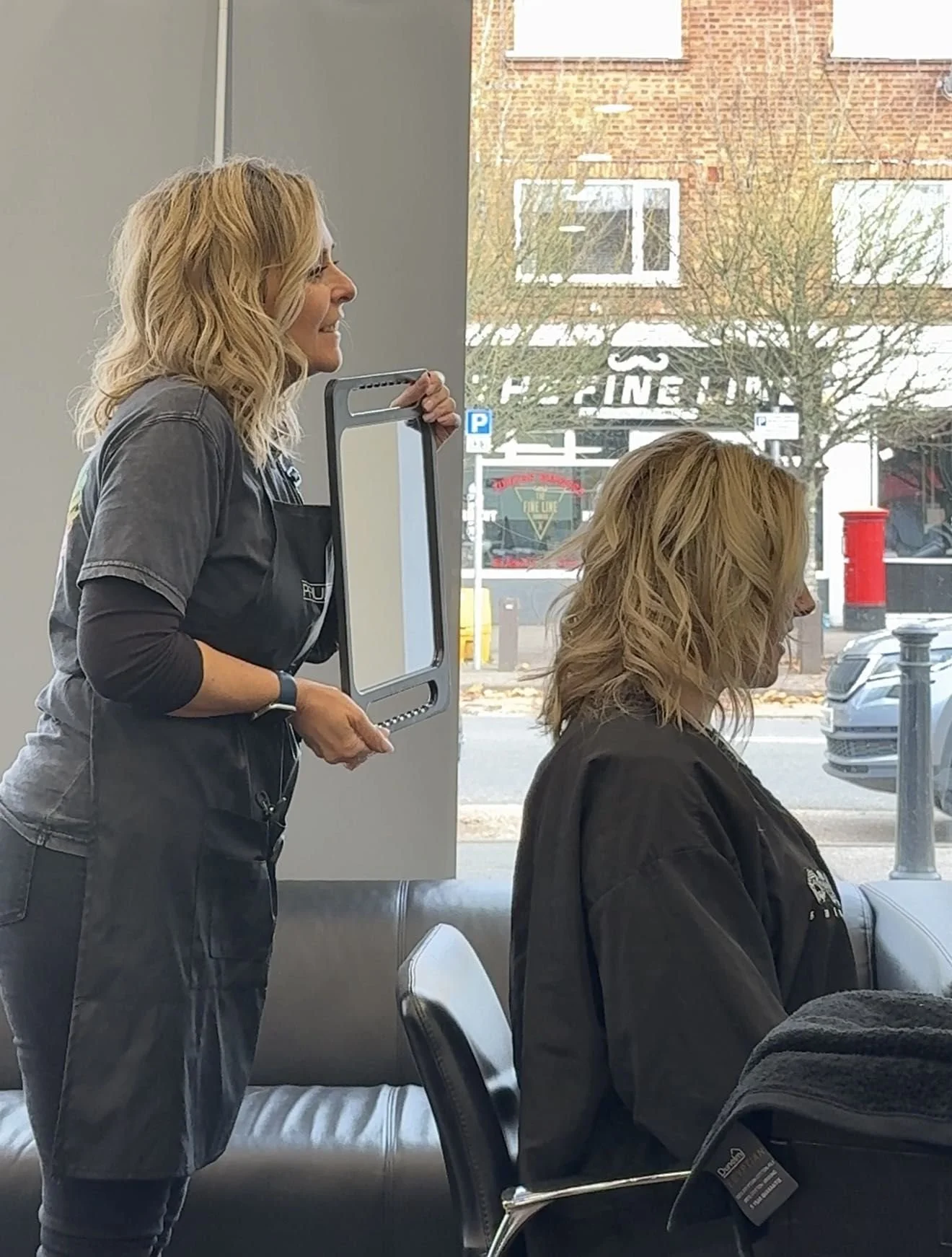 A woman sitting on a chair at a salon inside, with hair stylist standing behind her, and a large window showing an outside view of buildings, trees, a red mailbox, and parked cars.