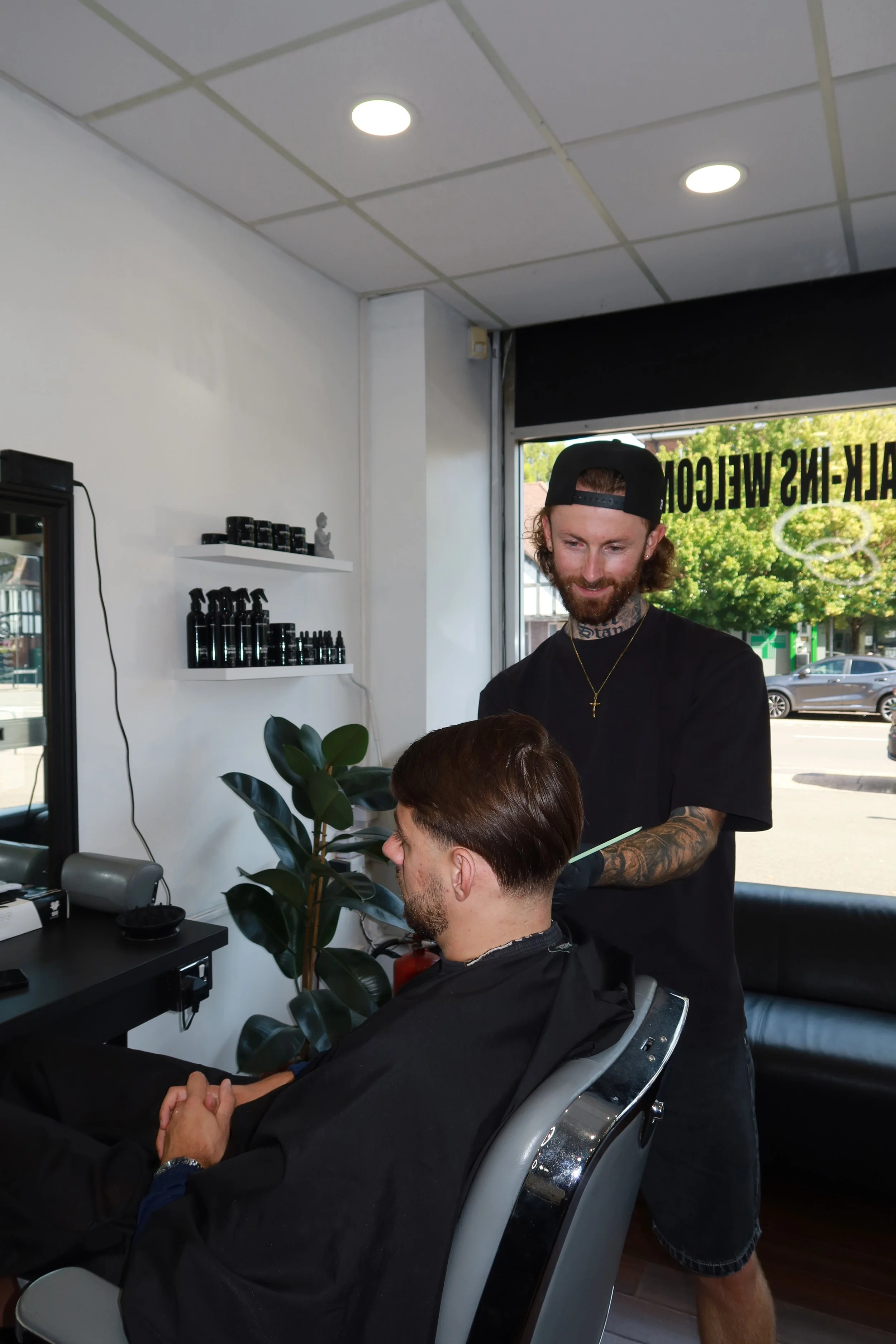 Barber trimming a young man's hair in a barbershop with white walls, ceiling lights, and various products on the counters.