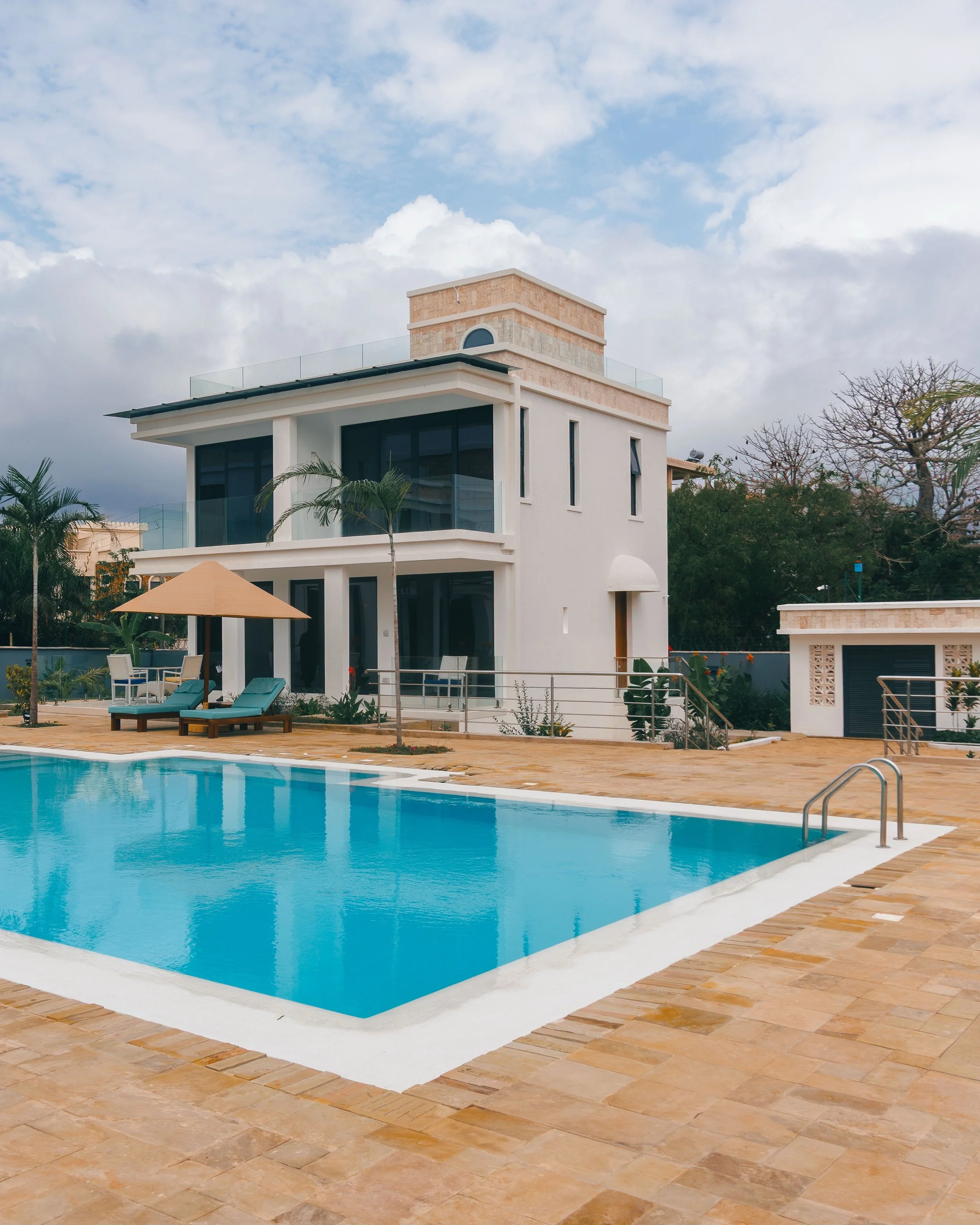 Lines Against the Coast &mdash; Amkia Villa, Diani

I kept coming back to the pool. The water sits still enough to hand you the whole house twice. Inside, a glass roof lays shadows down in neat bars, and three blown-glass pendants turn in the stairwe