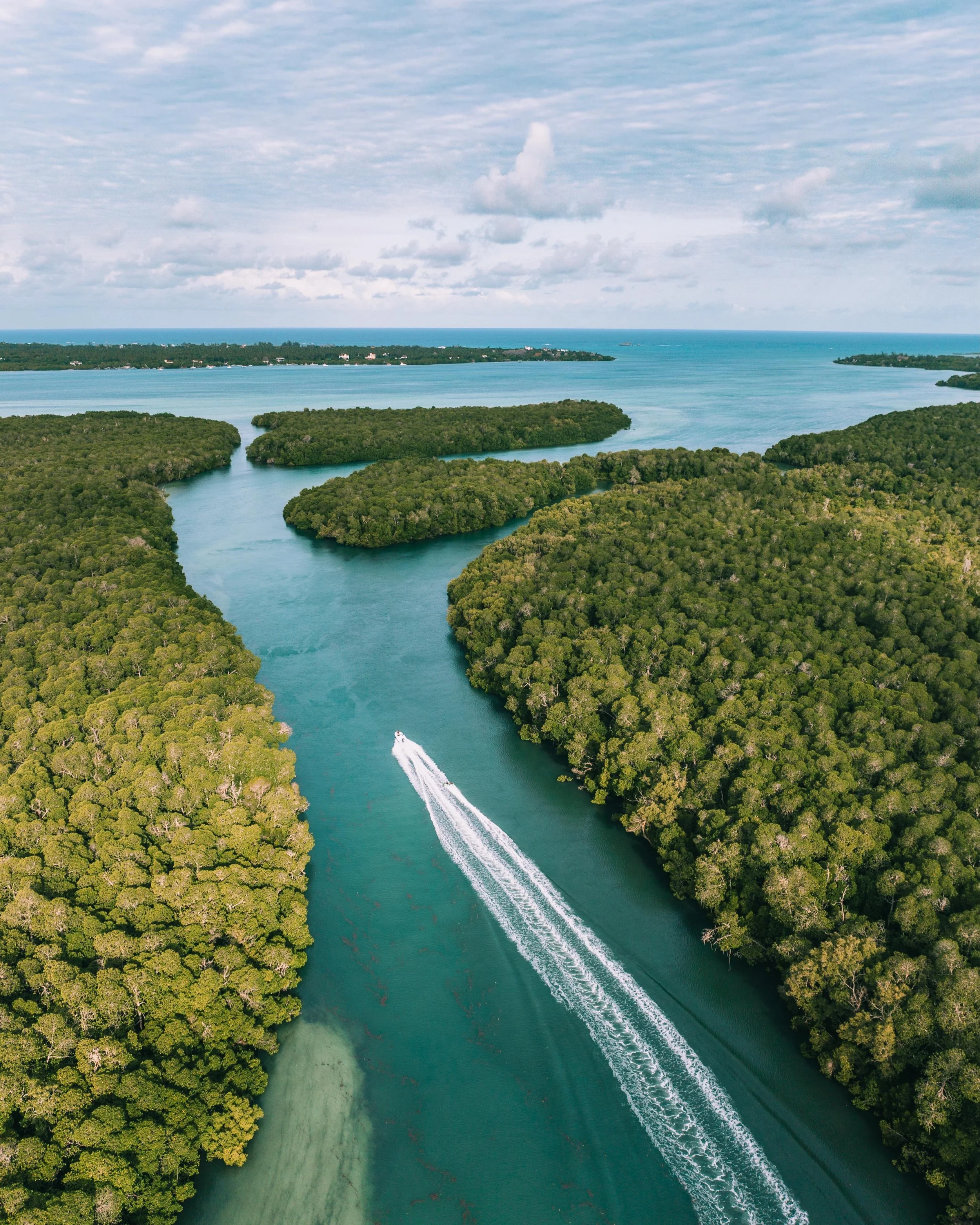Mida Creek opens up from above: mangrove islands stitched together by channels of jade water, a speedboat carving a white line through the middle. Behind it, the tag boat game pulls riders over the sandbanks, their wake splitting the shallows in two.