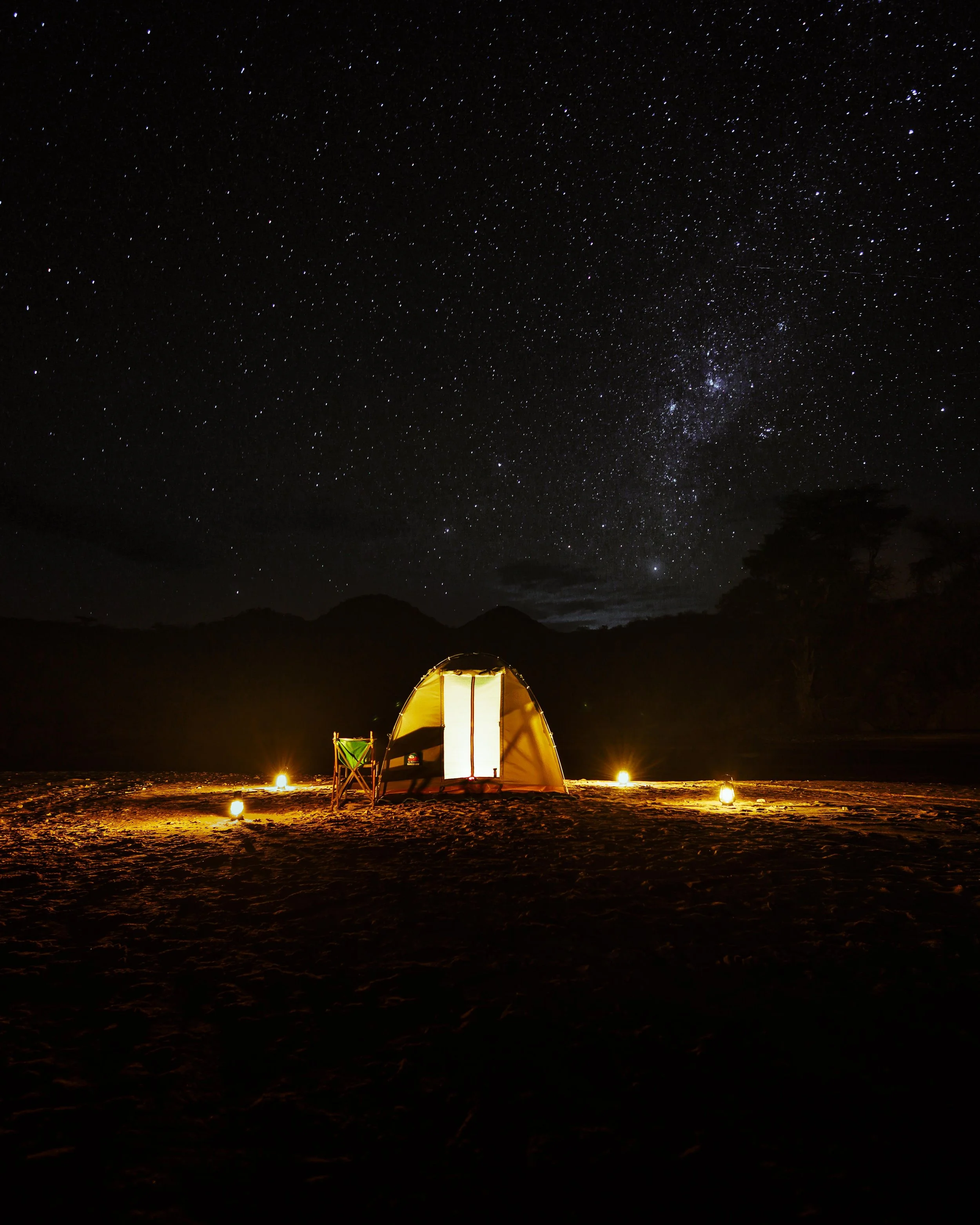 A nighttime scene with a starry sky above a camping tent illuminated from within. The tent is set up on rocky terrain with small lanterns around it, and mountains are visible in the distance.