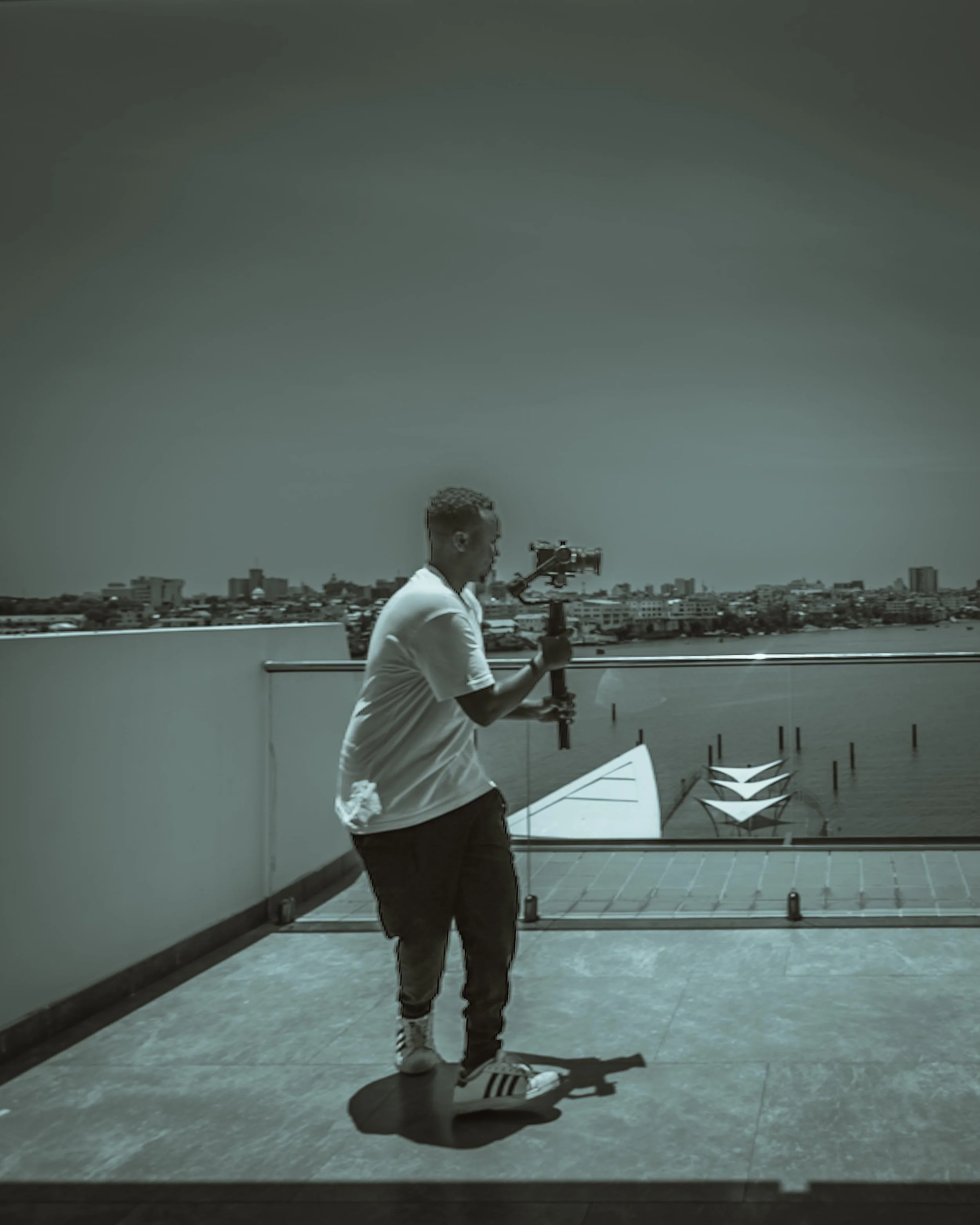 Person with short hair in white shirt standing on a rooftop with a city skyline in the background, holding a camera on a tripod, casting a shadow on the ground.
