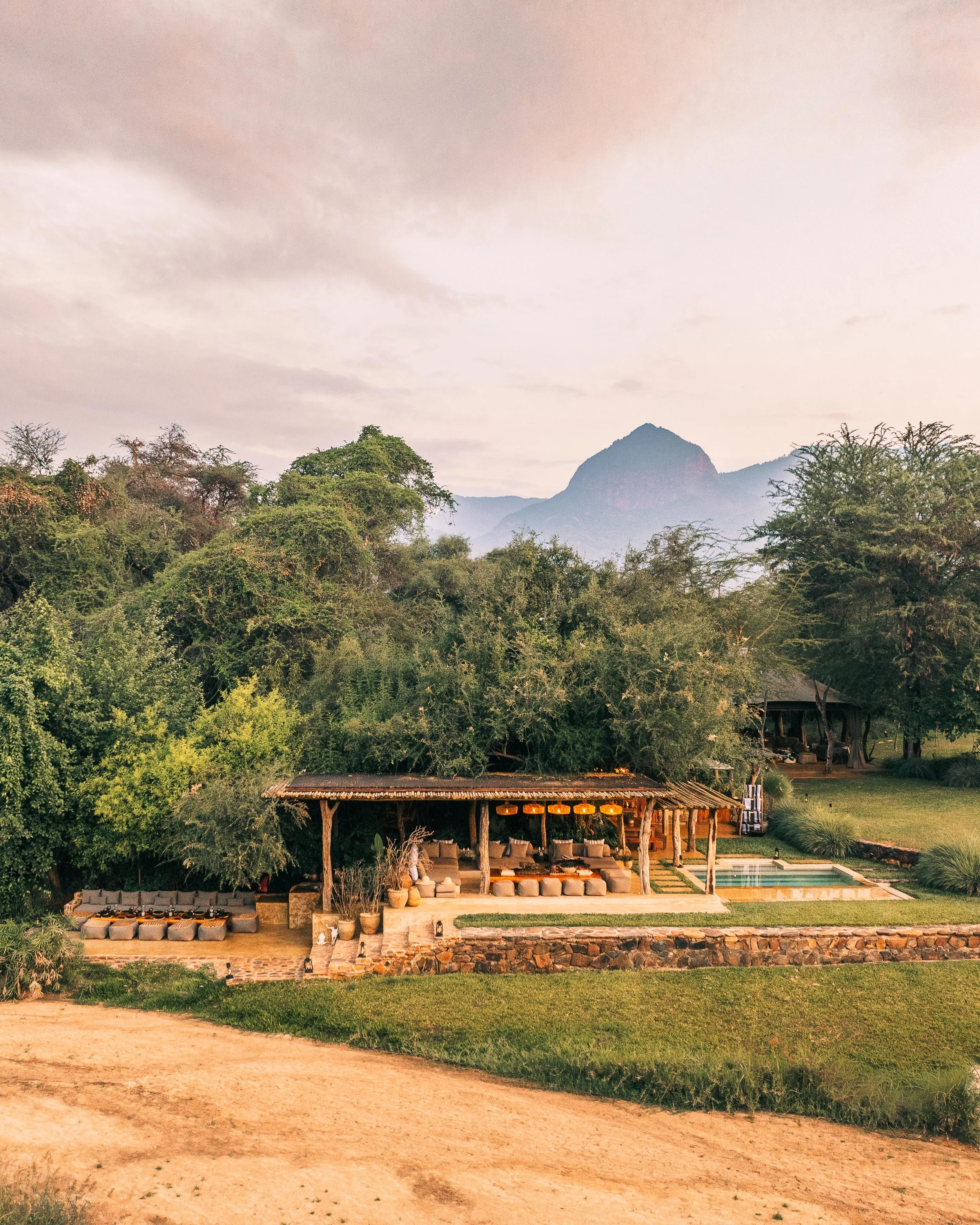 A tropical outdoor setting featuring a modern open-air lounge with beige couches, a swimming pool, lush trees, and mountains in the background under a cloudy sky.