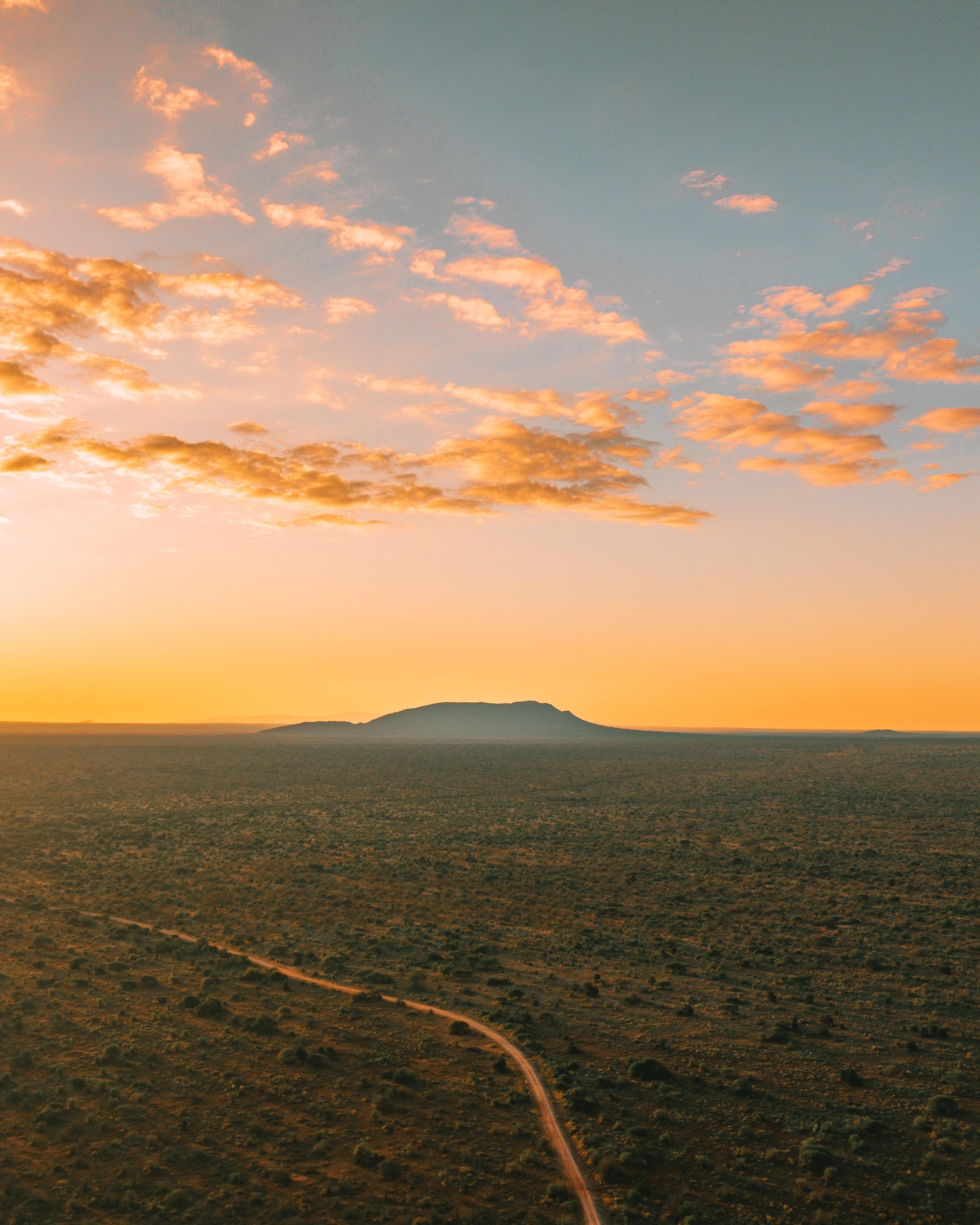 A sunset over a desert landscape with a winding dirt road and a mountain in the distance, clouds illuminated by the golden hour light.