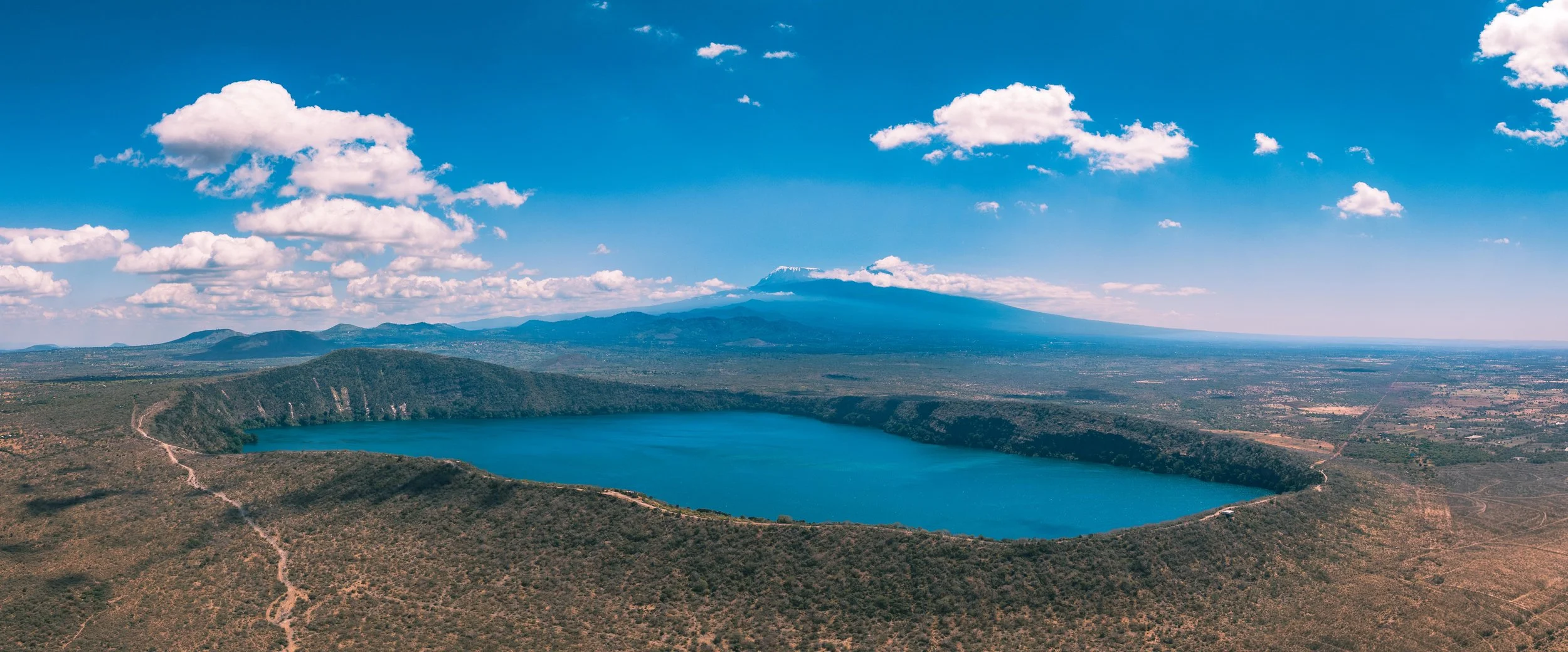 A large blue crater lake surrounded by green hills and mountains under a partly cloudy sky with Mount Teide in the background.