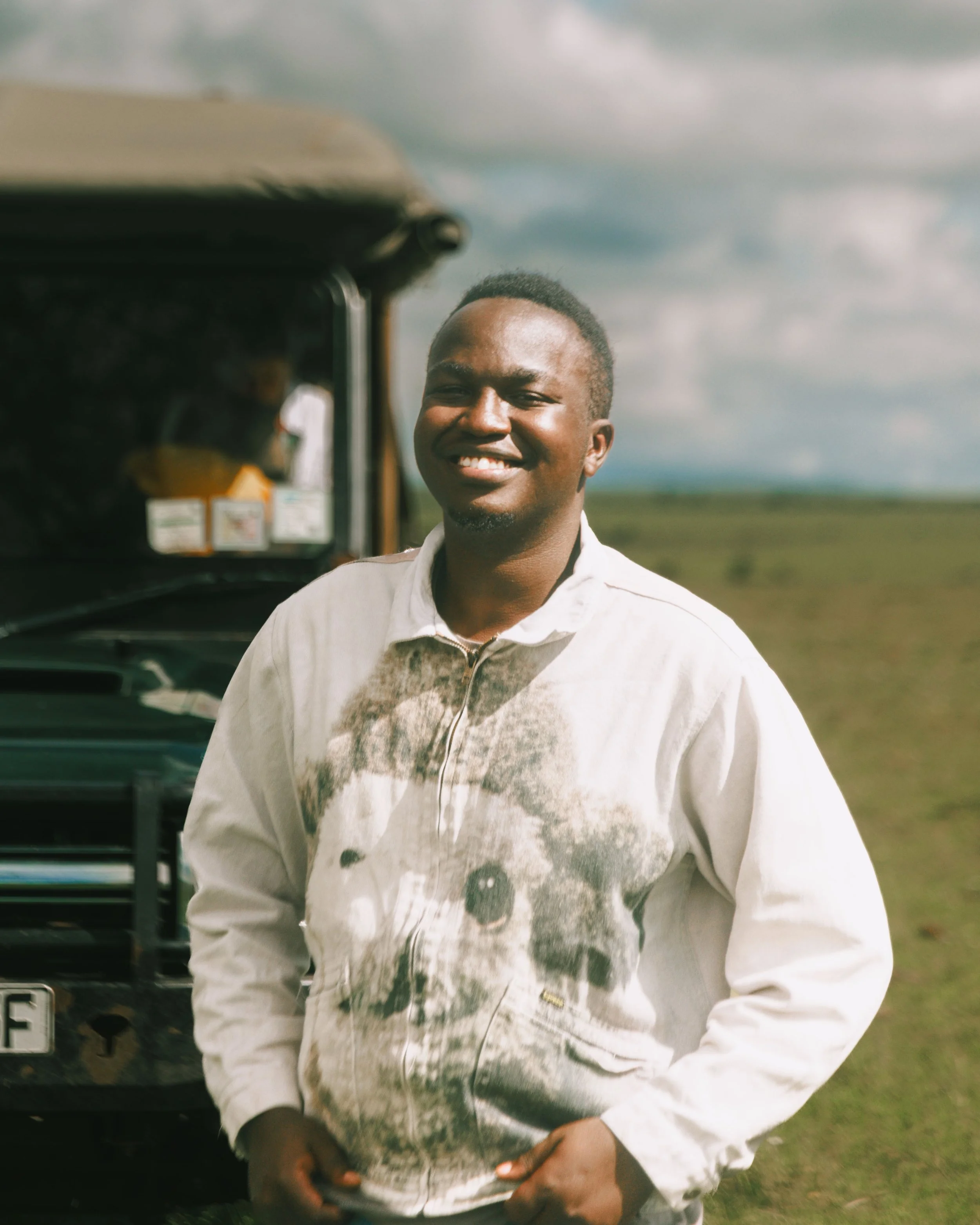 A young man with dark skin and short dark hair smiling outdoors, wearing a cream-colored jacket, with a grassy field and cloudy sky in the background.