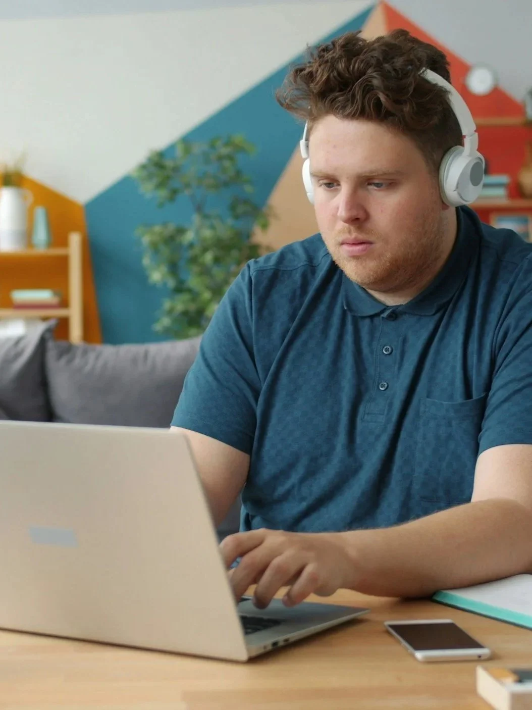 A young man wearing headphones working on a laptop at a wooden desk.