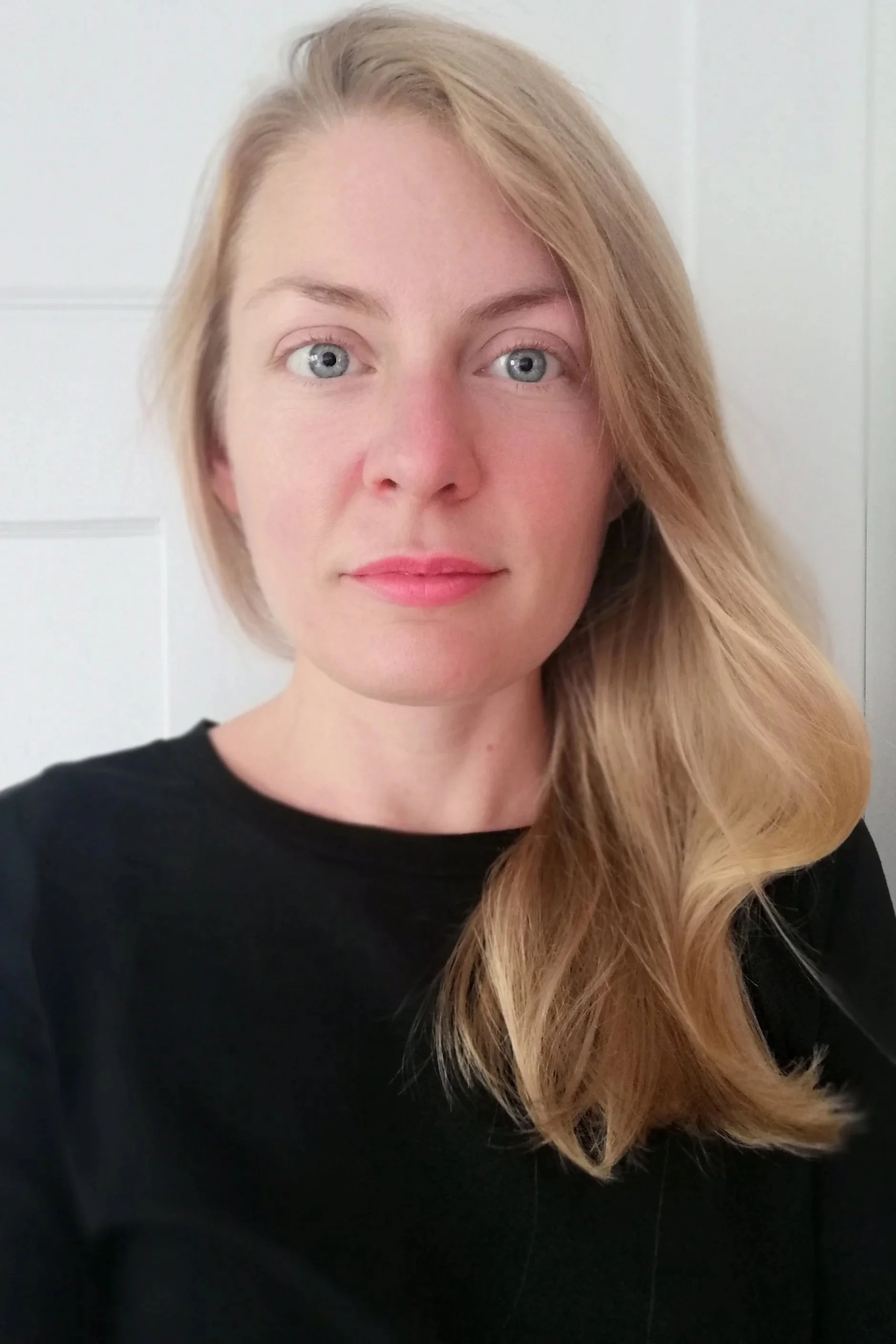A woman with blonde hair, blue eyes, and fair skin wearing a black top, standing against a white background.