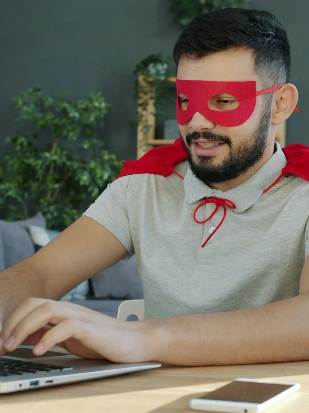 A man with a beard wearing a red superhero cape and a red eye mask, smiling while using a laptop at a desk.