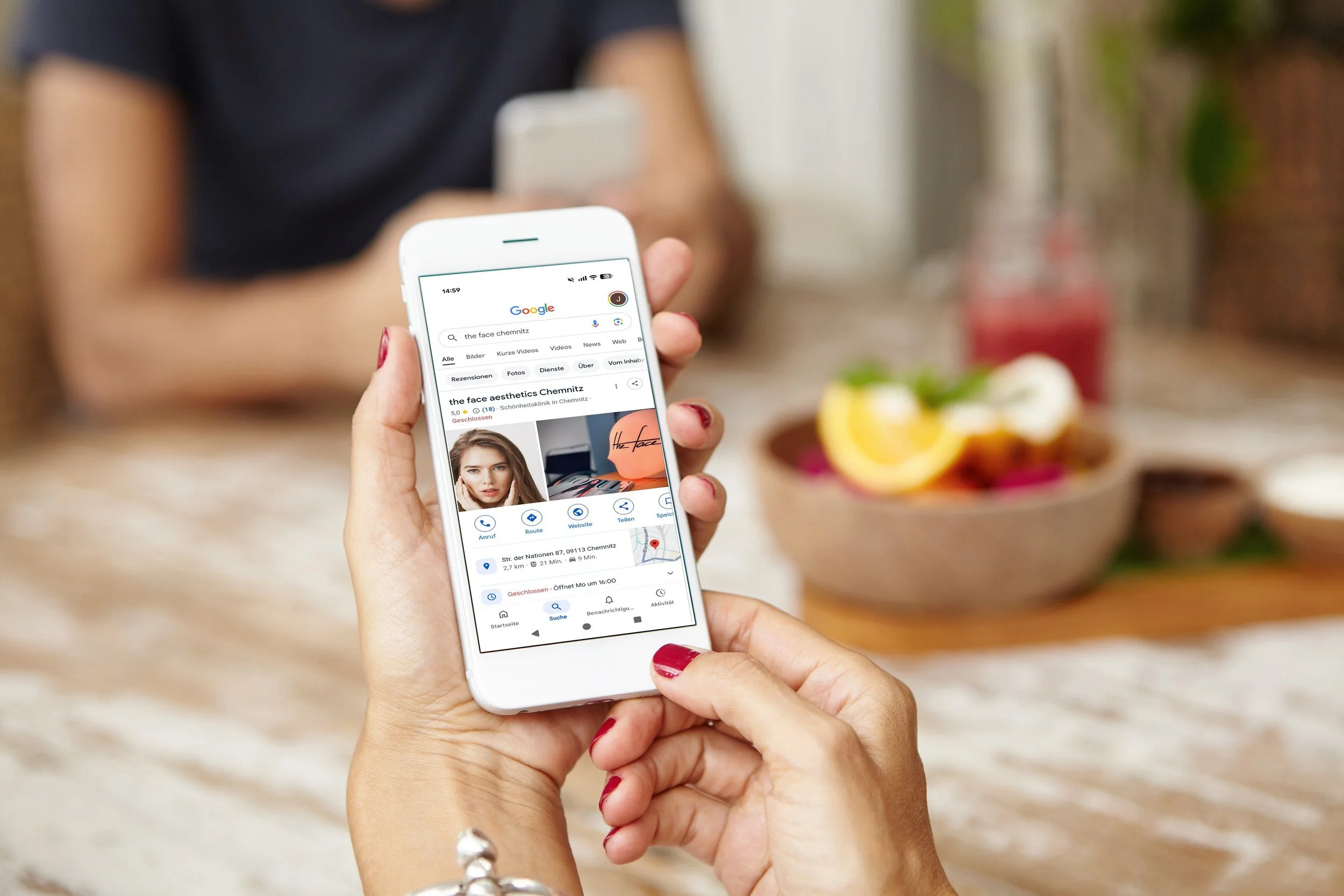 Person hält ein Smartphone mit Suchergebnis für die Praxis 'the face aesthetics Chemnitz' in der Hand, im Hintergrund eine Schüssel mit Obst und eine woonliche Umgebung.
