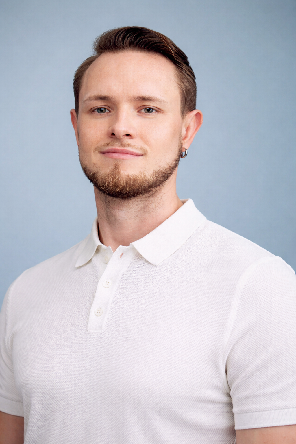 Junger Mann mit lila Augen, braunem Haar, Bart, Ohrring, trägt ein weißes Polo-Shirt, lächelt vor hellem blauen Hintergrund.