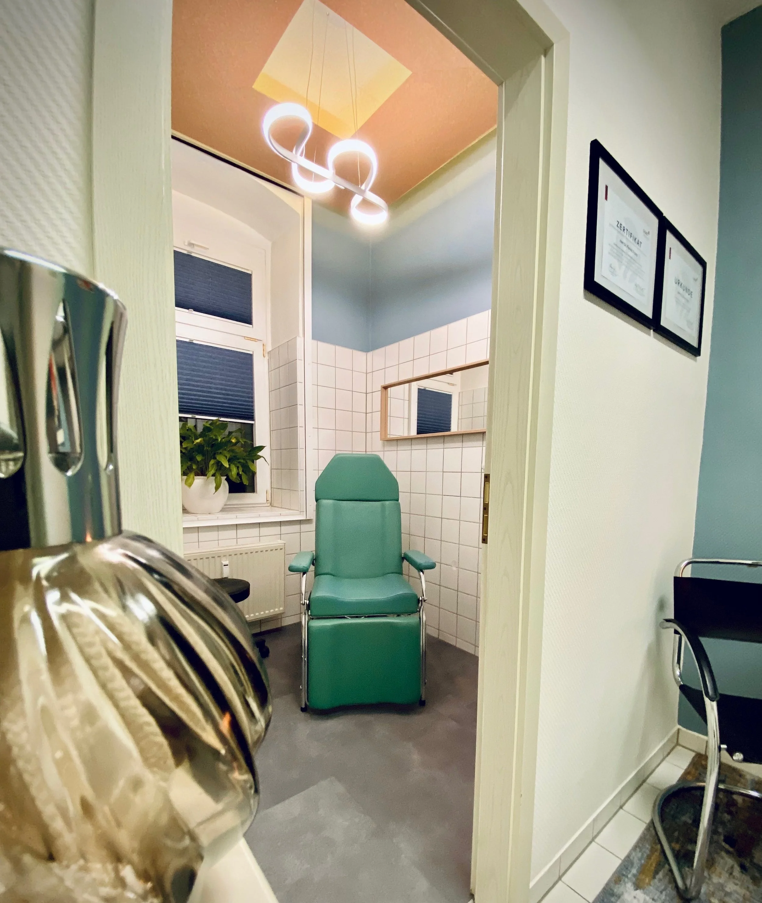 Eine Arztpraxis oder Behandlungszimmer mit einer grünen Klinikliege, einem Fenster mit Pflanze, weißen Fliesen an der Wand und moderner Beleuchtung an der Decke.