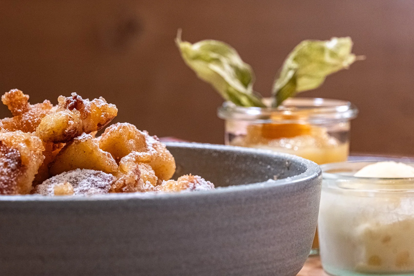 Frittierte Apfelchips in einer Schüssel mit Puderzucker, im Hintergrund Kräuter in einem Glas und zwei kleine Gläser mit Marmelade und Vanillepudding.