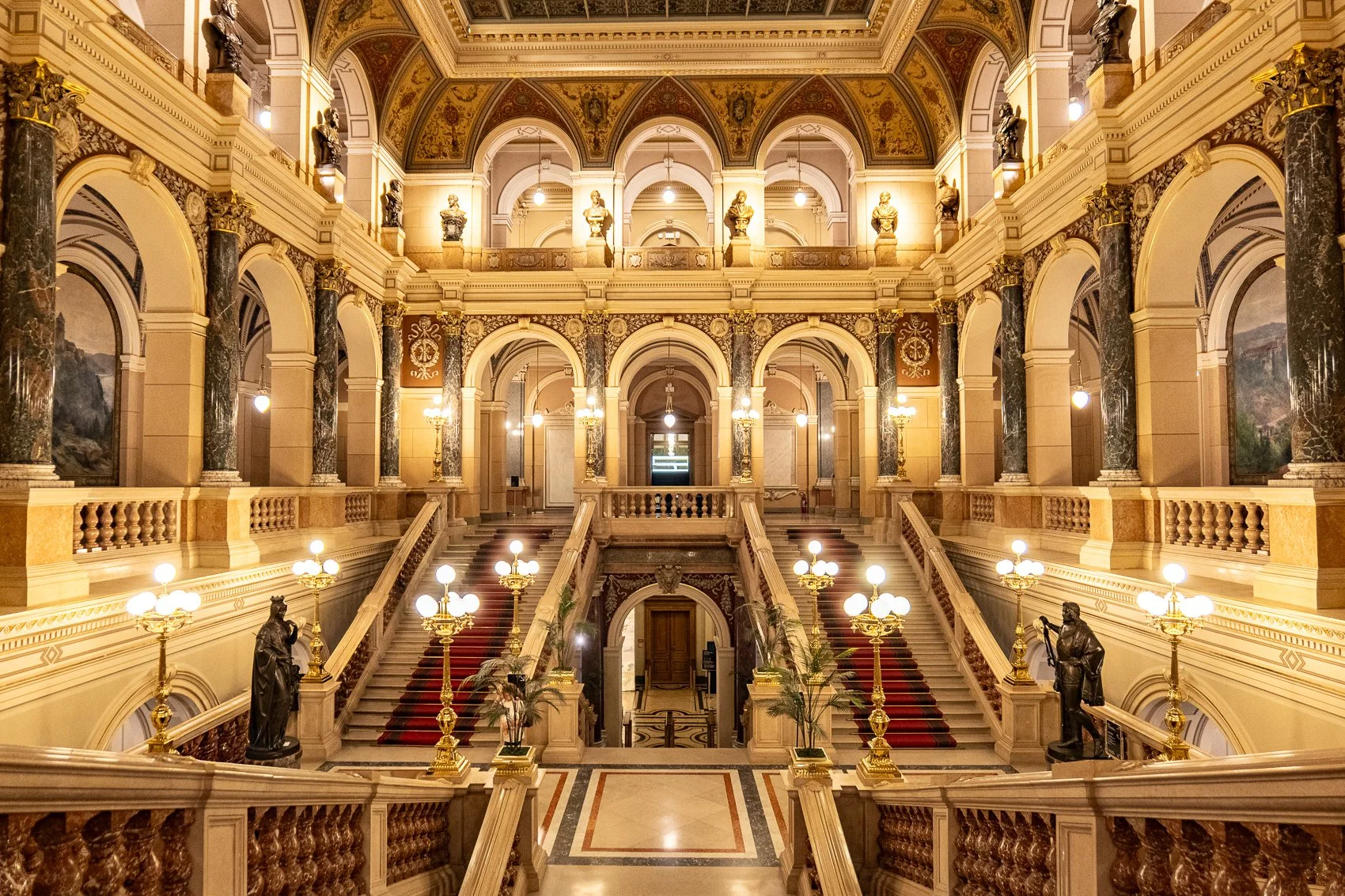 Ein prunkvoller, eleganter Innenraum mit Treppen, Säulen und Statuen im Rittersaalstil, beleuchtet durch mehrere antike Lampen. Der Raum hat eine reich verzierte Decke und dekorative Wandmalereien.