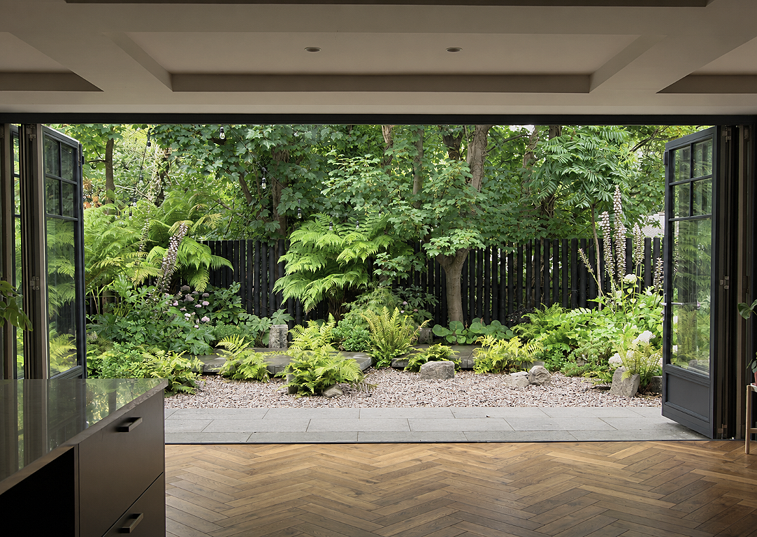 View of a lush green garden with various plants, ferns, and trees seen through open sliding glass doors from an indoor space with wooden flooring.